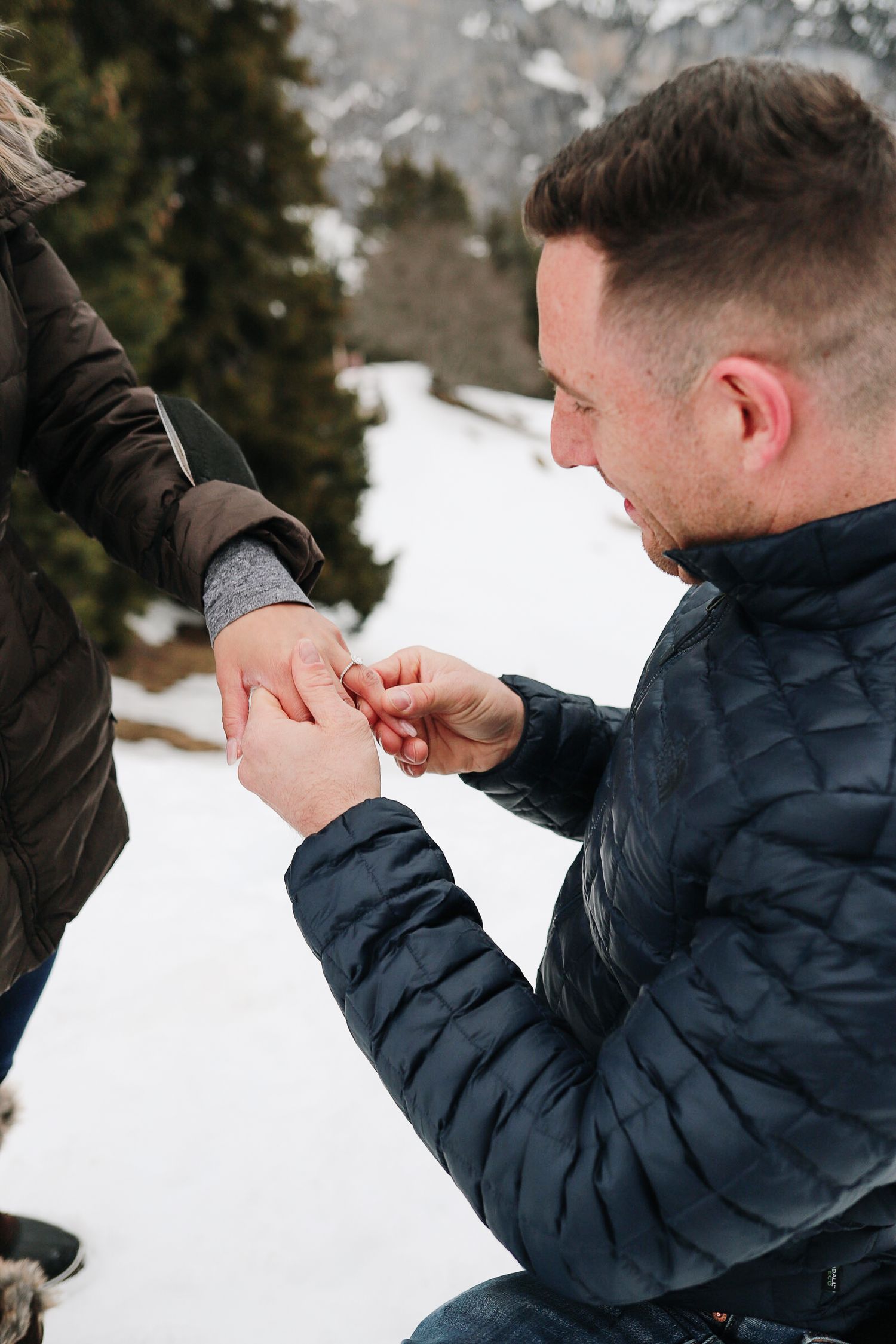 Man kneeling in snowy landscape placing engagement ring on womans hand during outdoor winter marriage proposal.