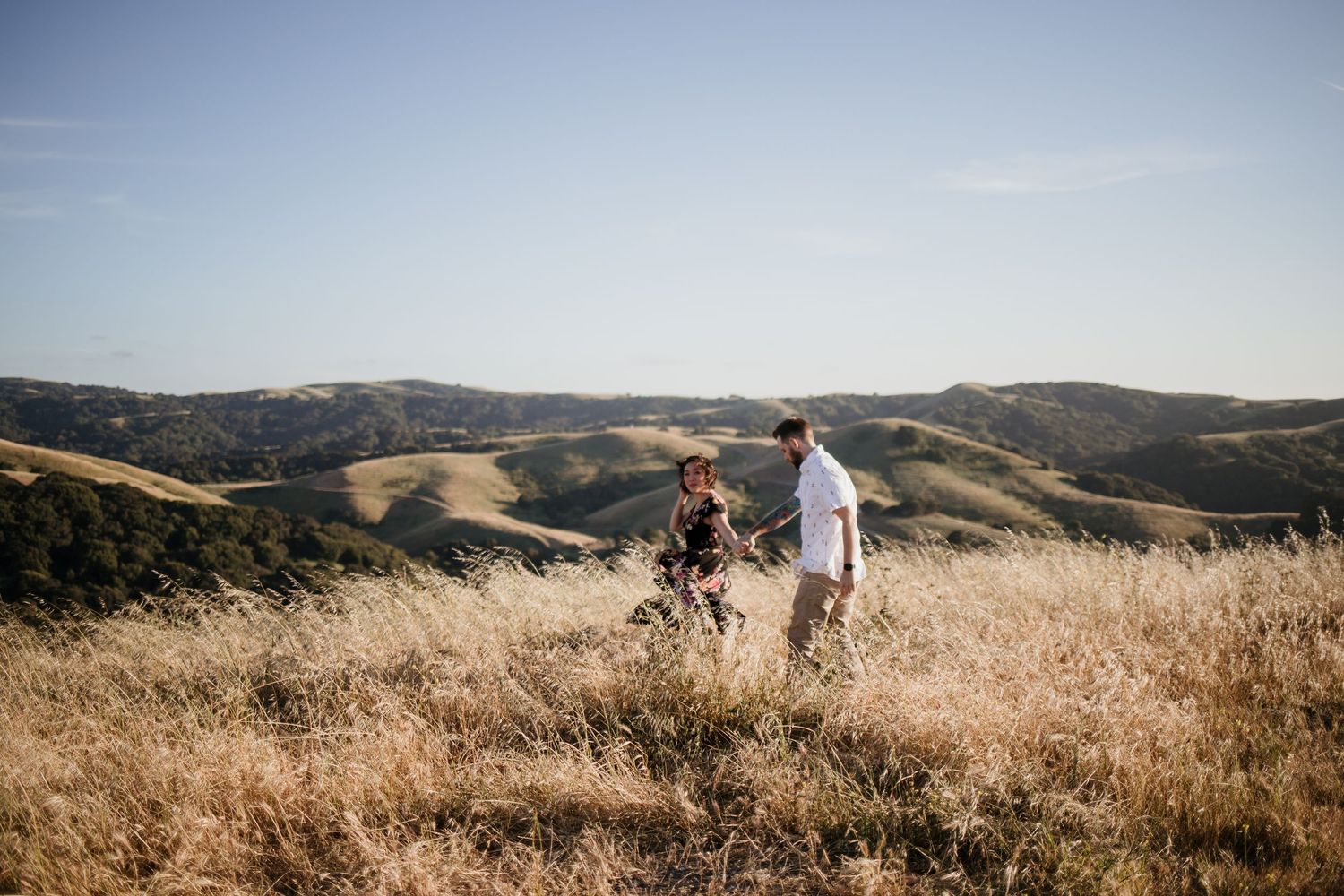 Couple walking through golden hillside meadow with rolling green mountains and clear blue sky backdrop in Dublin, California.