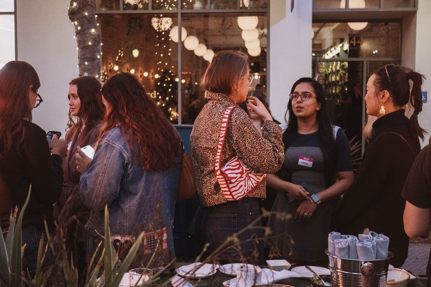 Women networking at an indoor evening social event with warm string lights and lush greenery in the background.