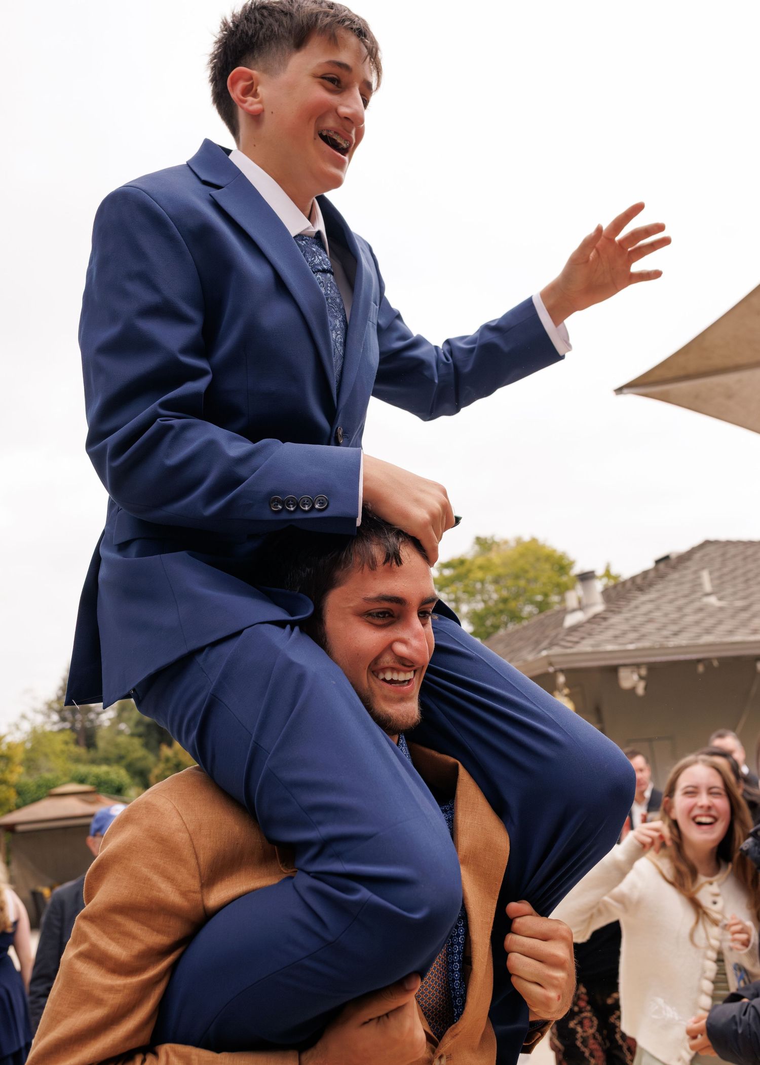Two young men in blue suits celebrate outdoors, one riding on the other's shoulders, laughing at a formal event.