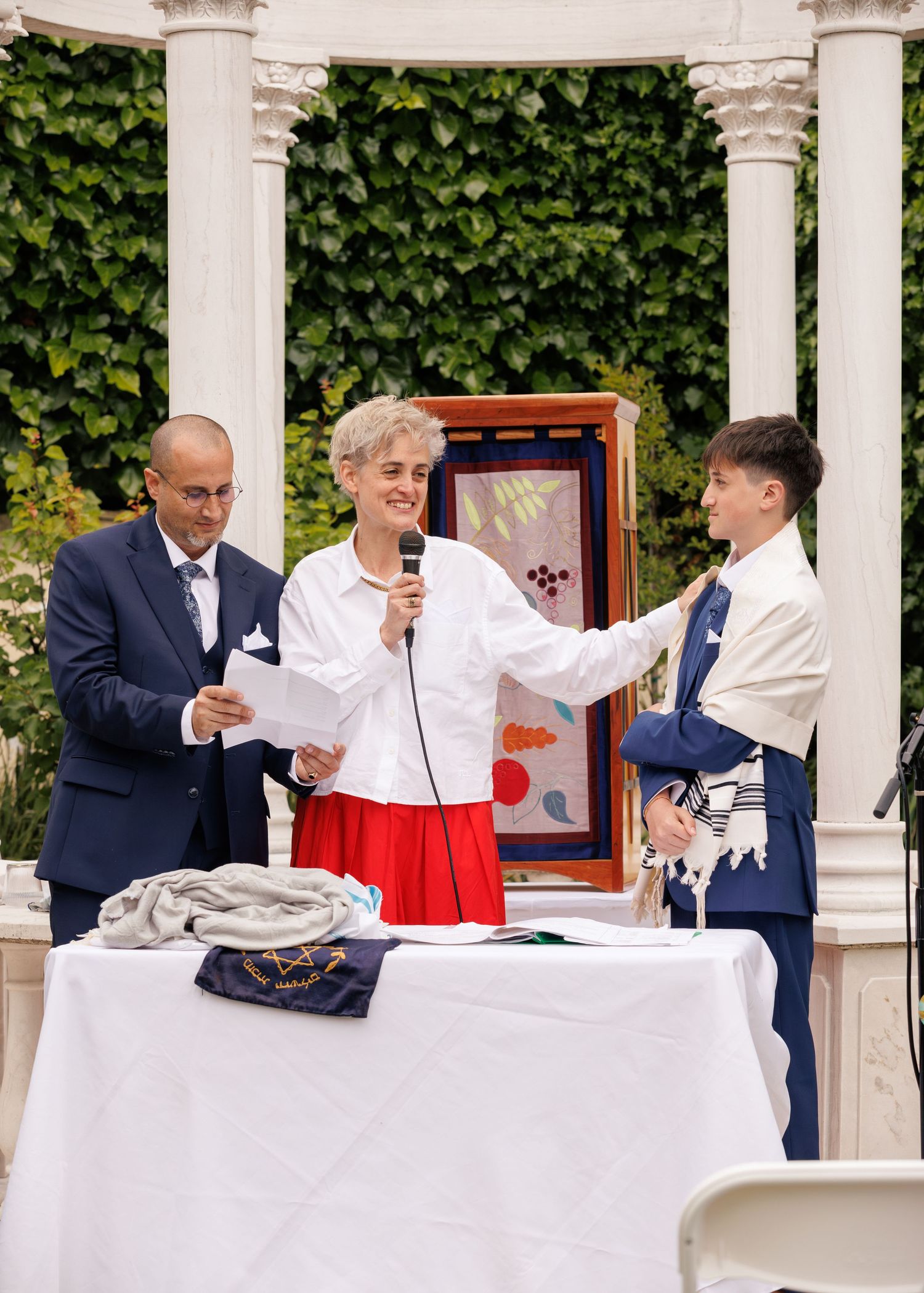 Three people stand at a white-draped table during an outdoor Bar Mitzvah ceremony beneath a white gazebo.