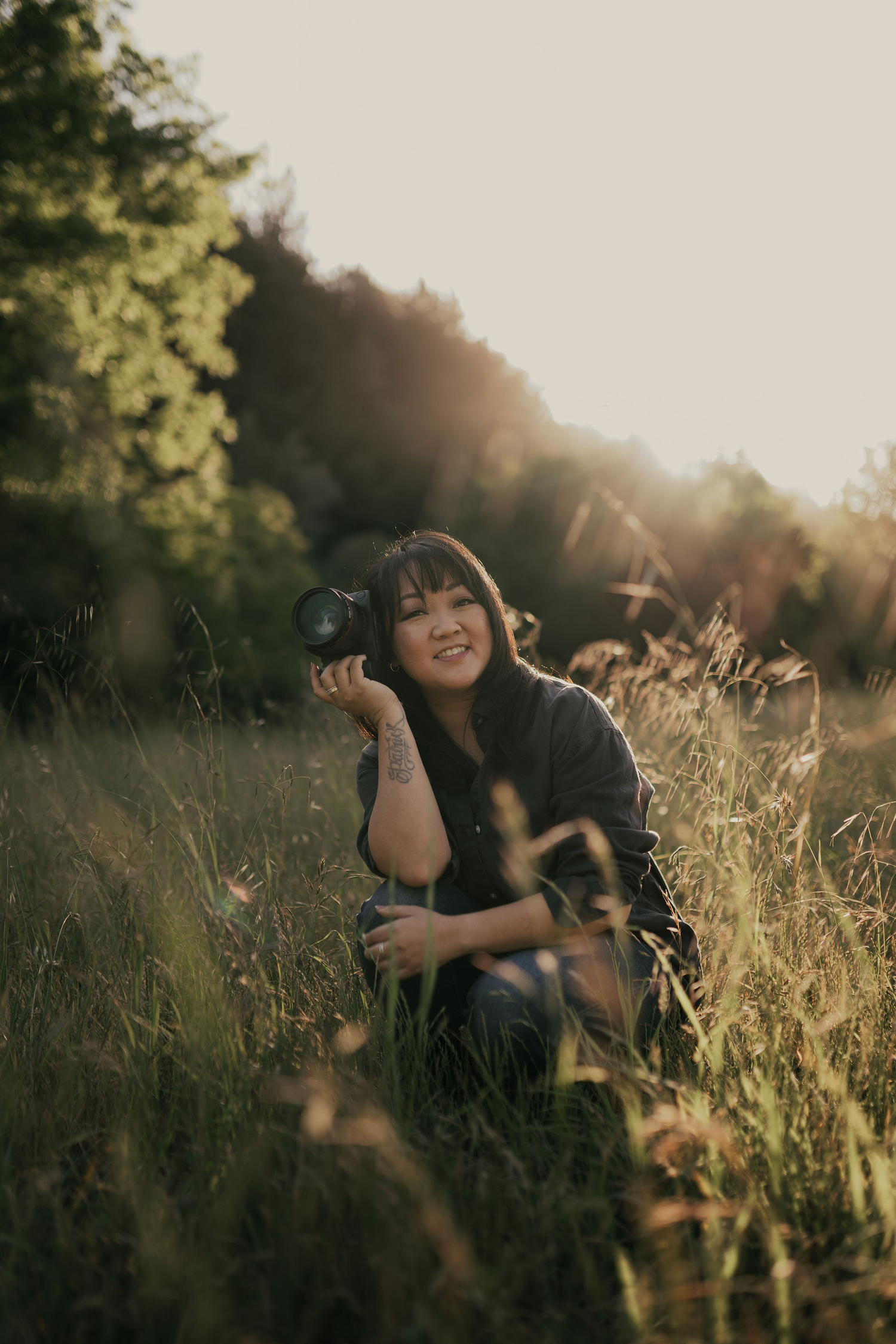 Photographer with camera sits in golden sunlit meadow of tall grass during sunset.