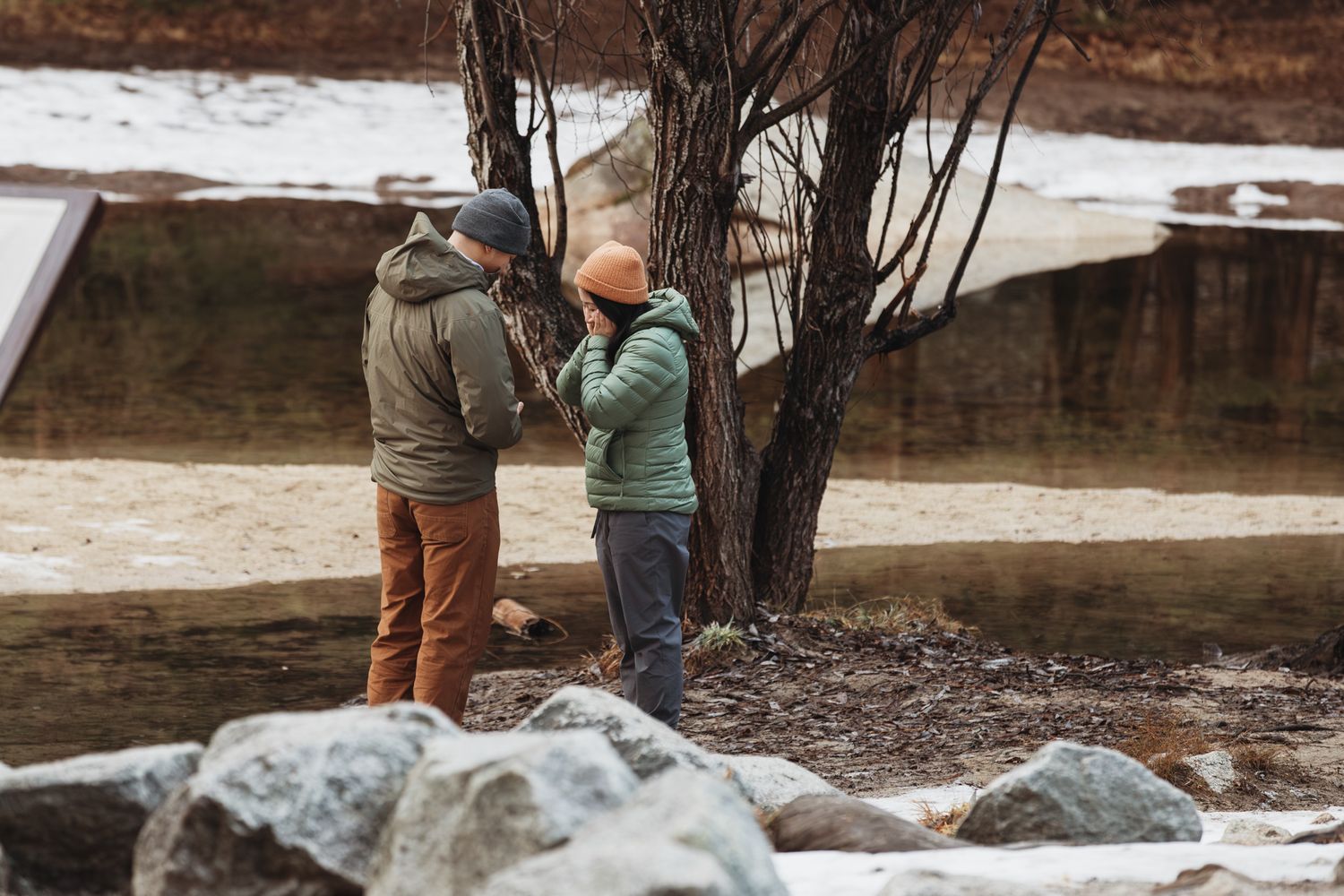 Two people in winter jackets stand near a bare tree by a snowy riverbank surrounded by rocks.
