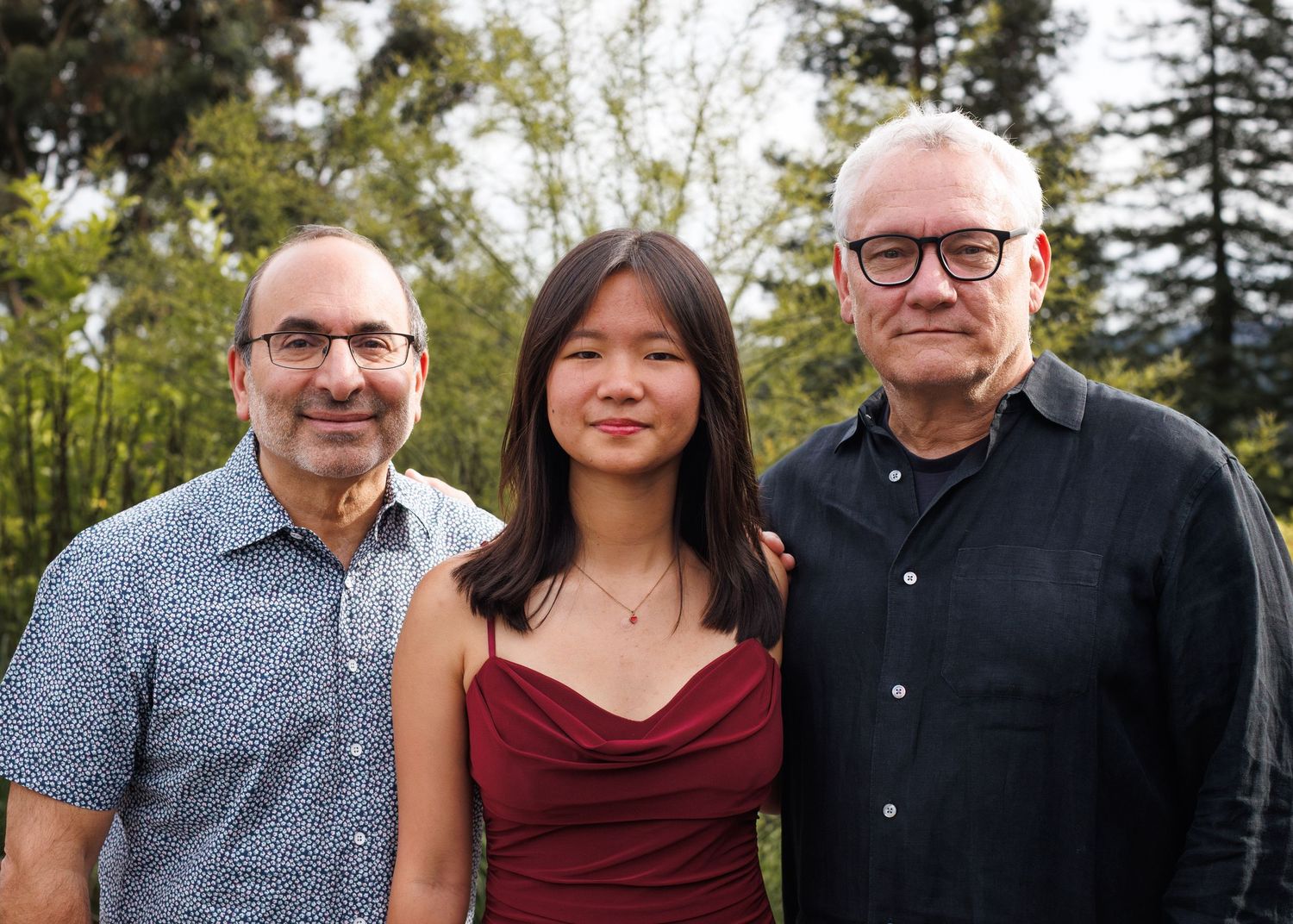 Three people smiling outdoors among trees, two older men flanking a young woman in a red dress.