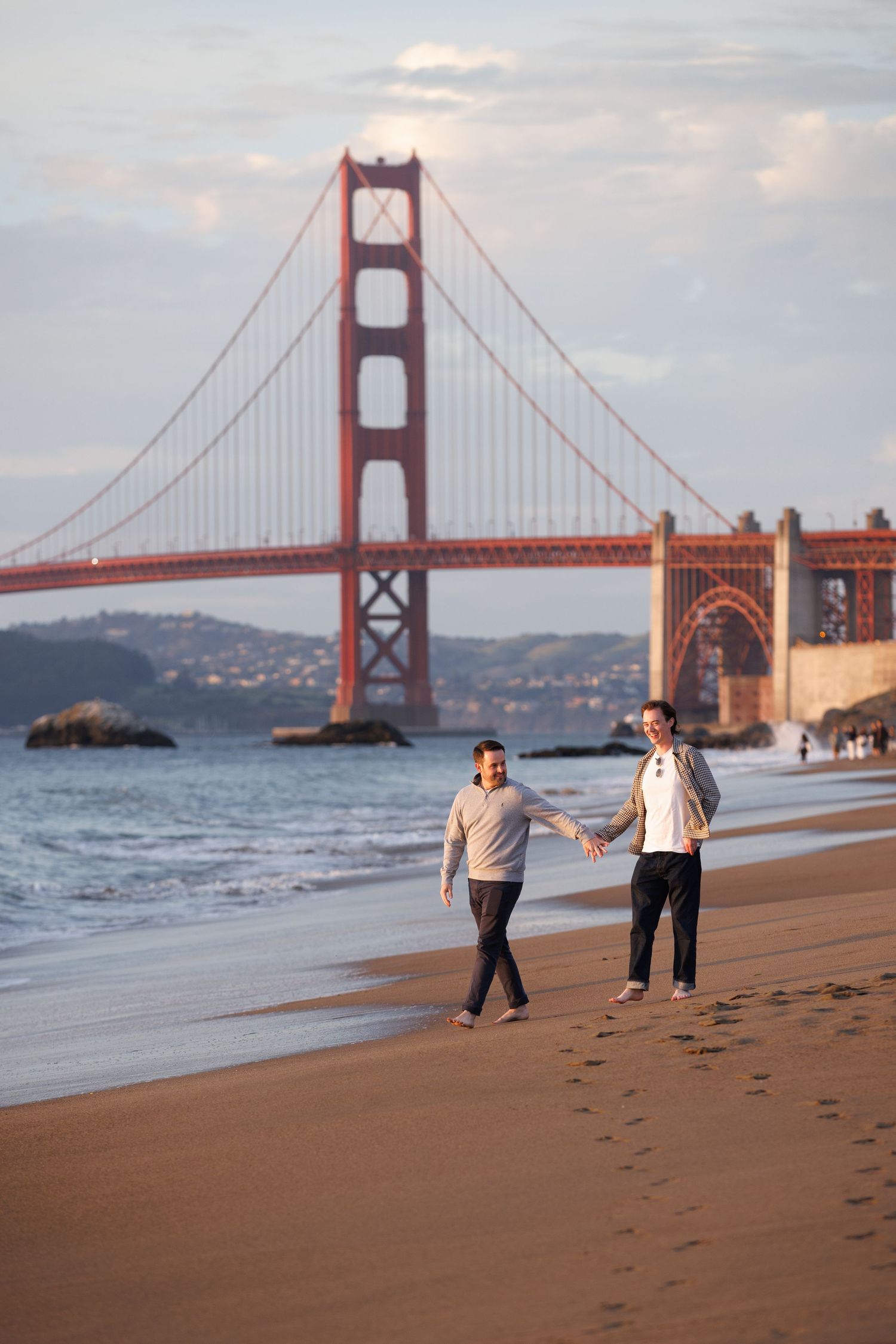 Couple holding hands walking on Baker Beach with the iconic Golden Gate Bridge in the background at sunset.