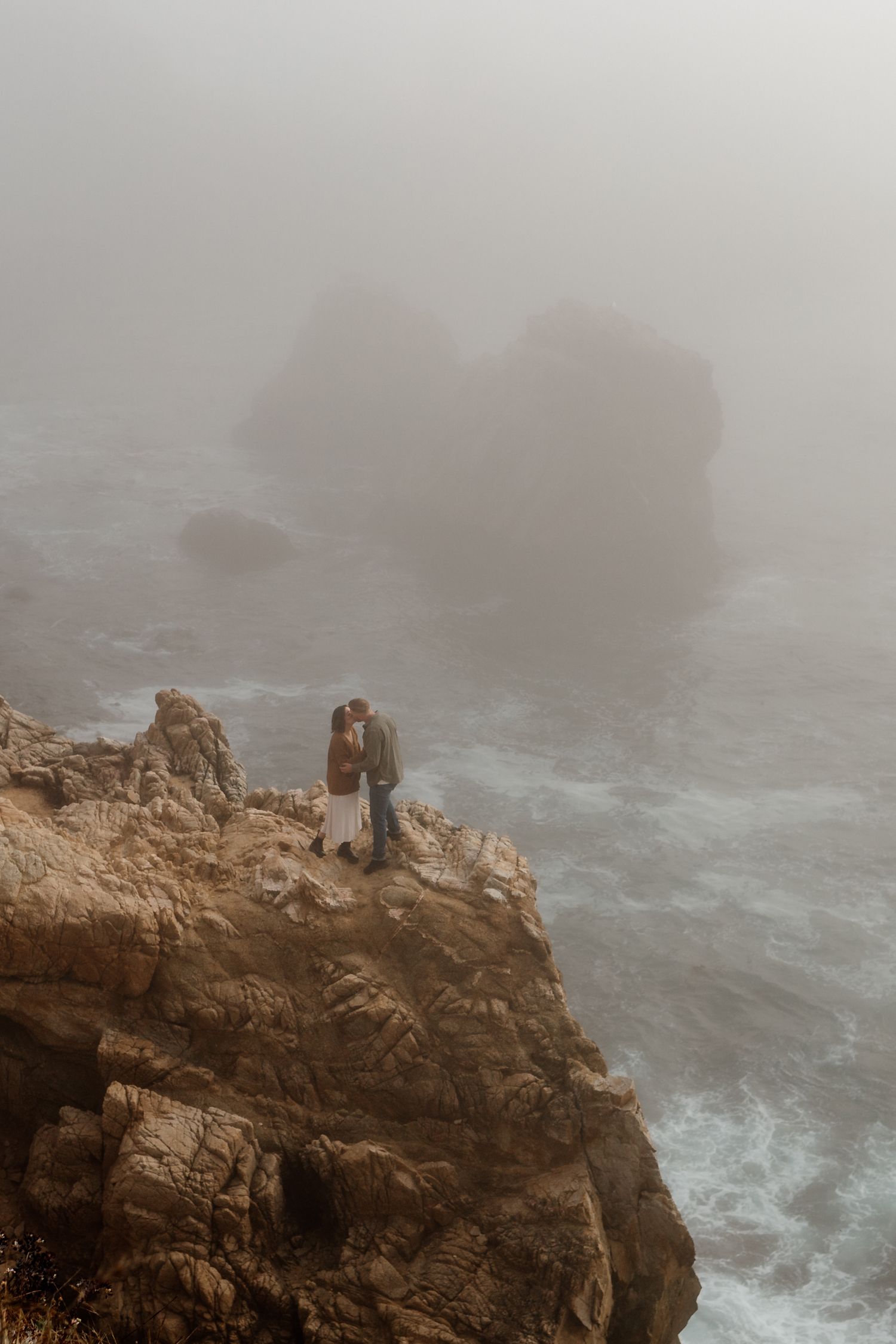 Couple embracing on rocky coastal cliff in misty fog with churning ocean waves below.