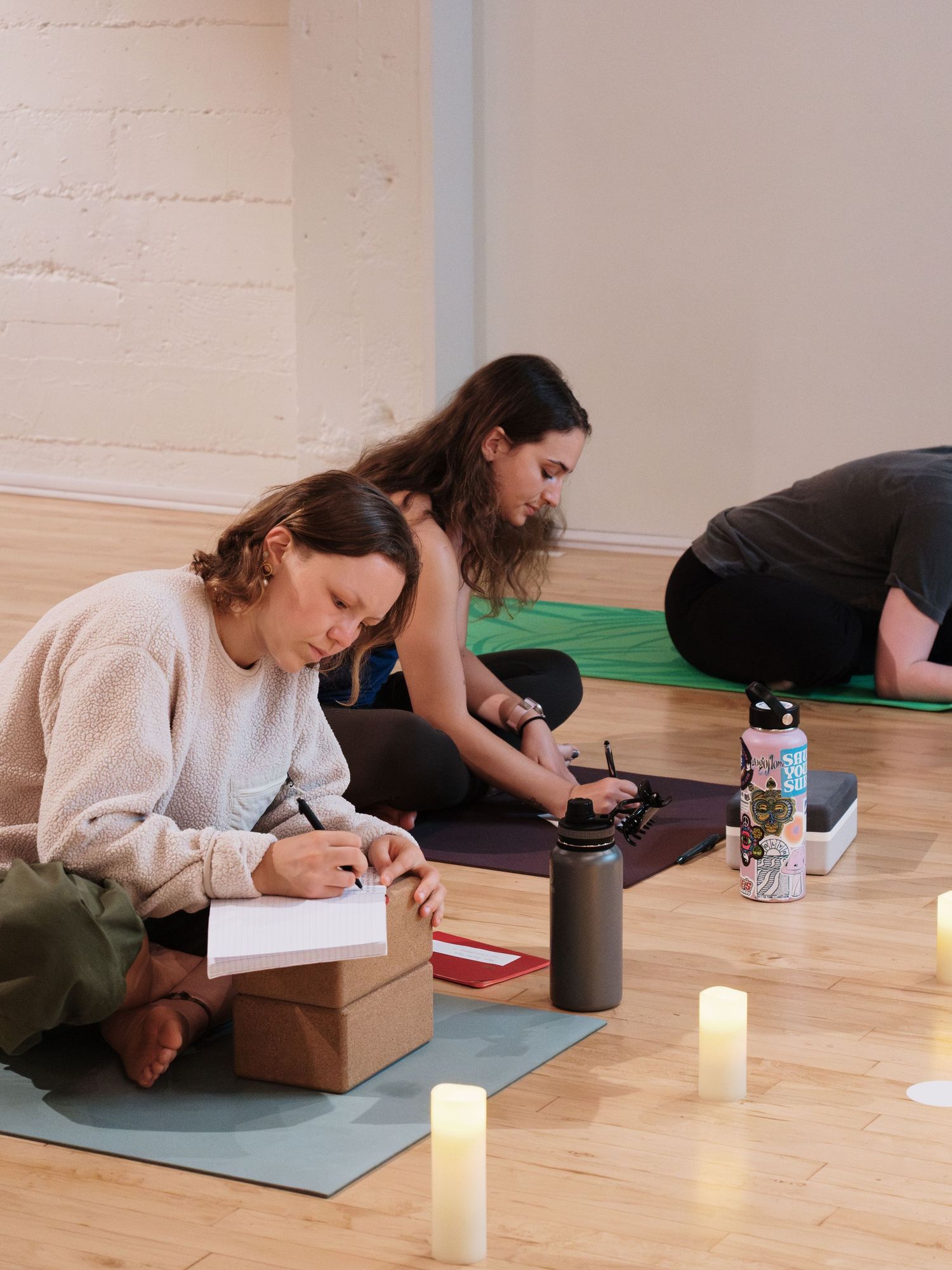 Women sitting on yoga mats with candles writing in notebooks during a candlelit yoga journaling session.
