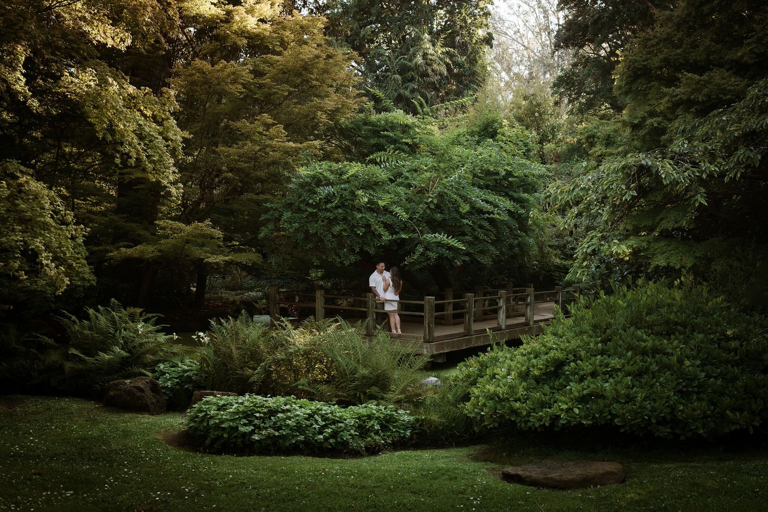 Couple embracing on a wooden bridge surrounded by lush green trees and gardens in a serene Japanese-style park.