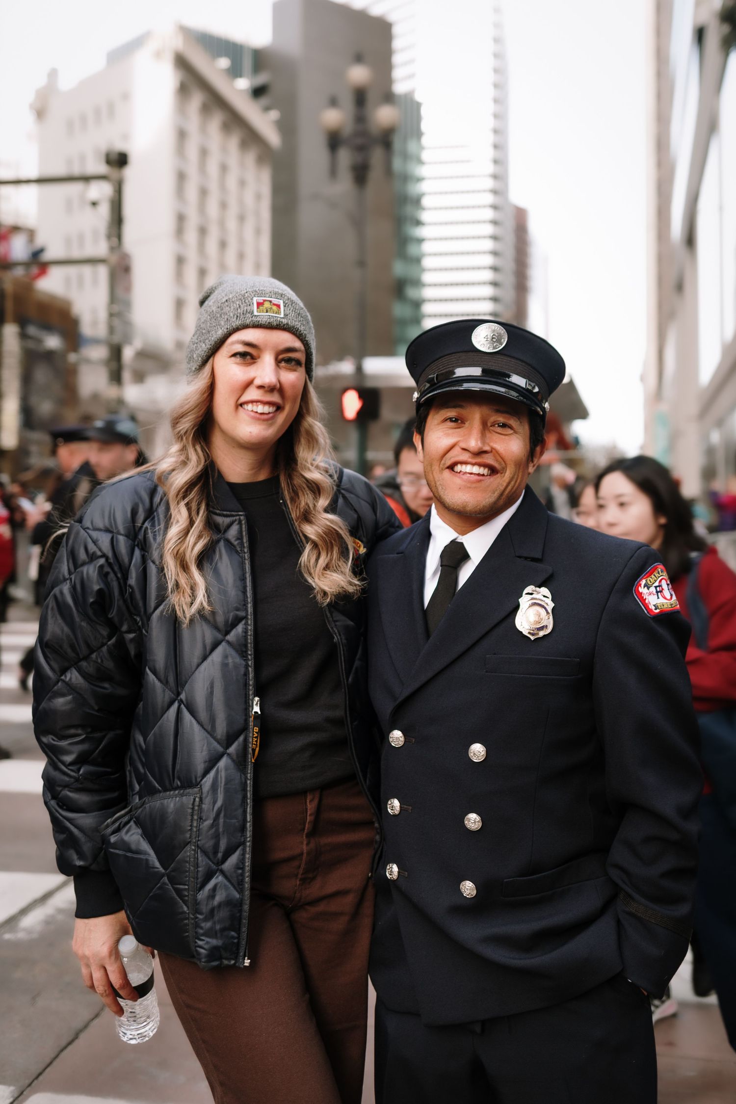 A woman in a beanie and jacket poses with a firefighter in uniform on a busy city street.
