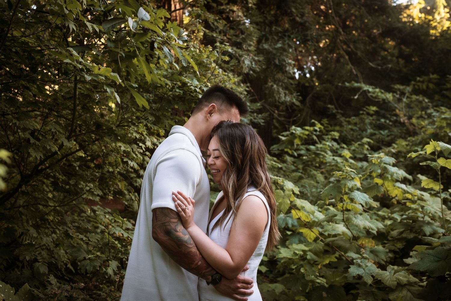 A couple embraces tenderly in a lush green forest, sharing an intimate moment surrounded by trees.