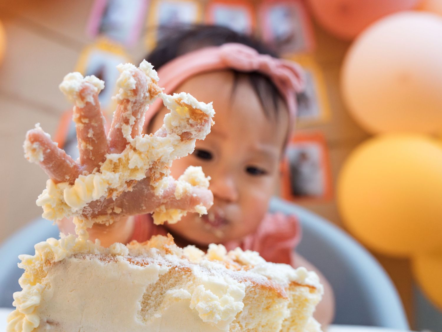 Baby girl with pink headband smashes her first birthday cake, covering her hand in white frosting.