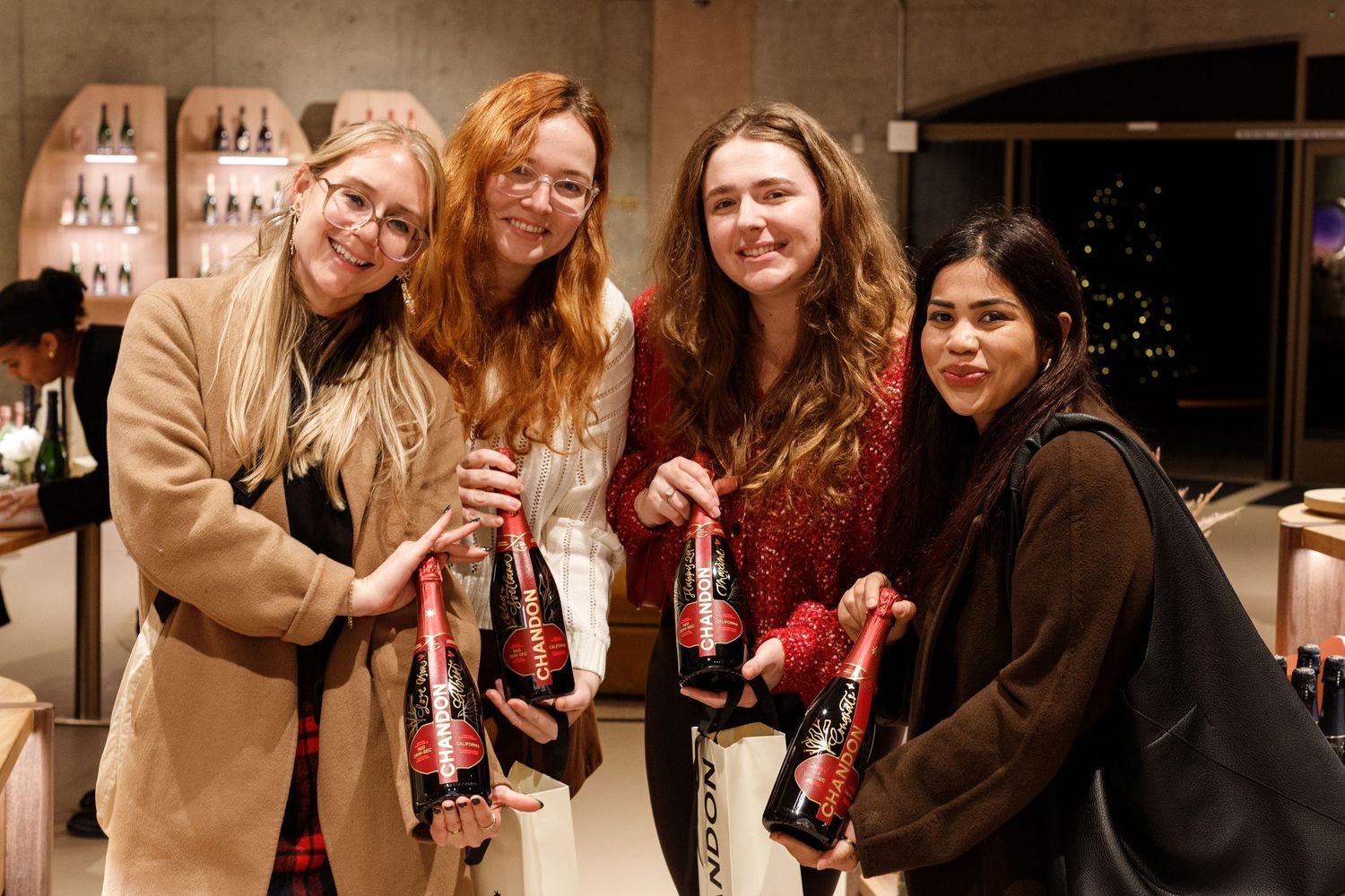 Four women smiling and holding red-labeled bottles at an indoor holiday party event.