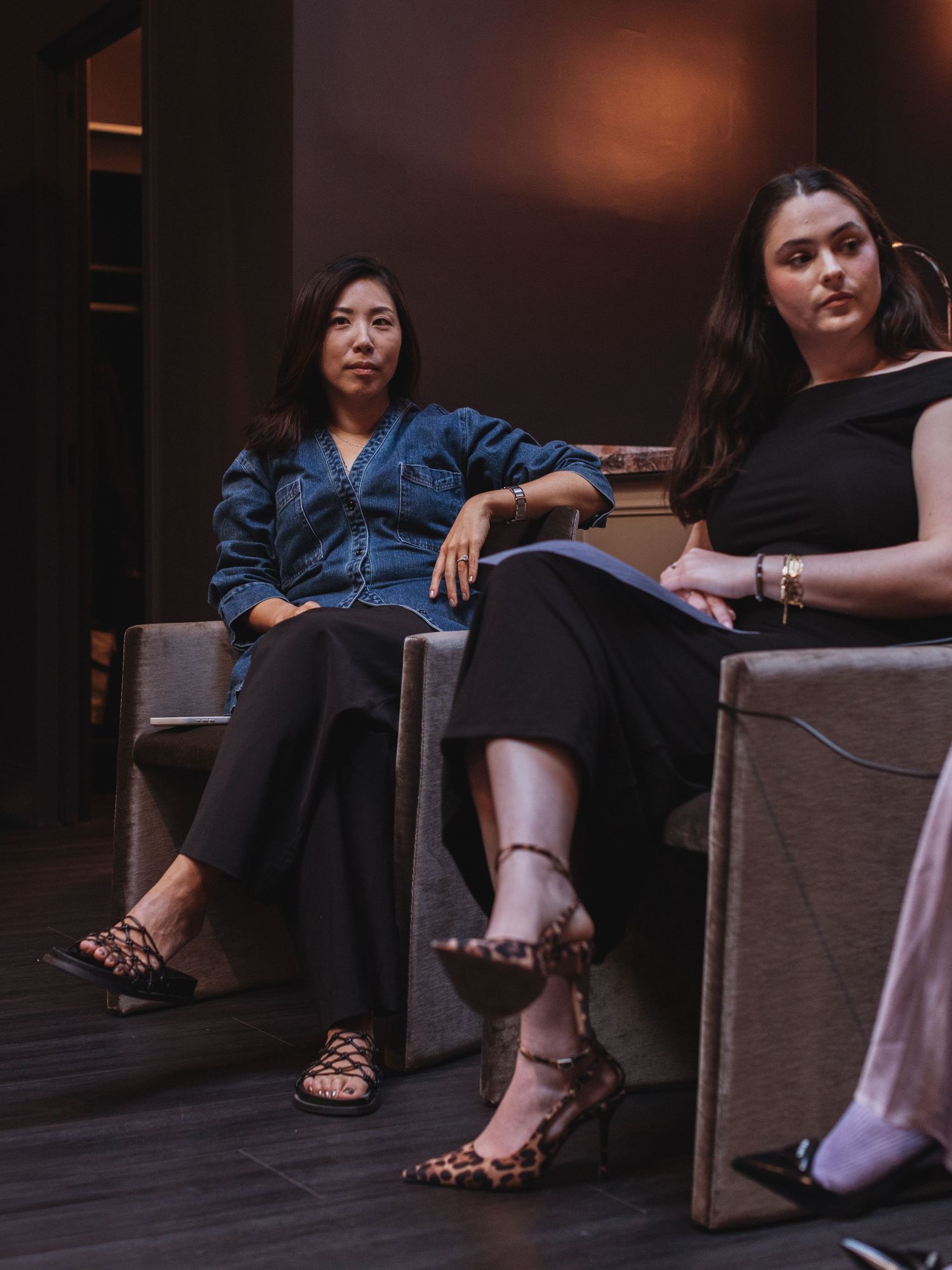 Two women in black outfits and leopard print heels sit in chairs during a panel discussion in a dimly lit room.