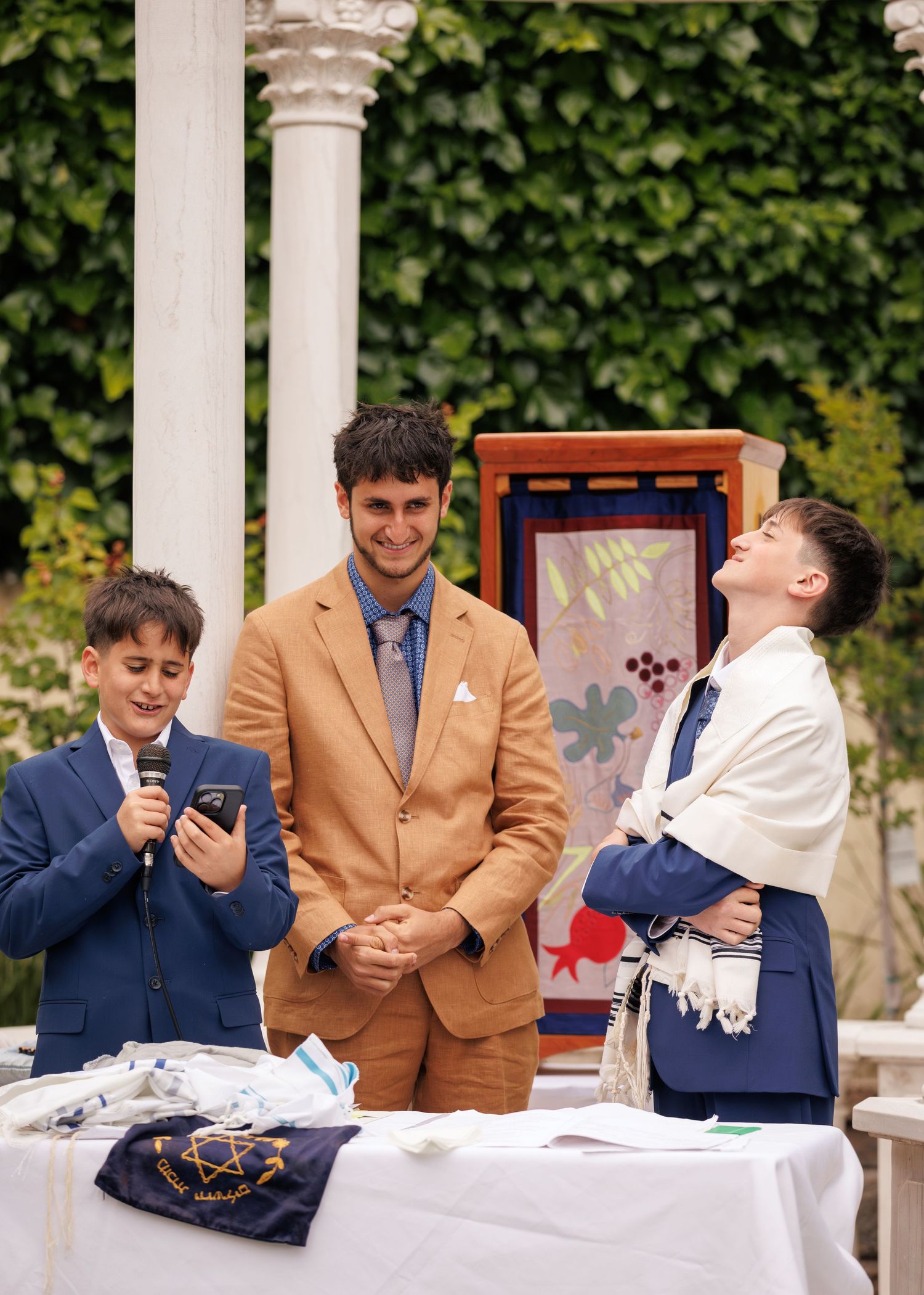 Three boys in formal attire at a Bar Mitzvah ceremony, one wearing a tallit and reading from a phone at an outdoor venue.