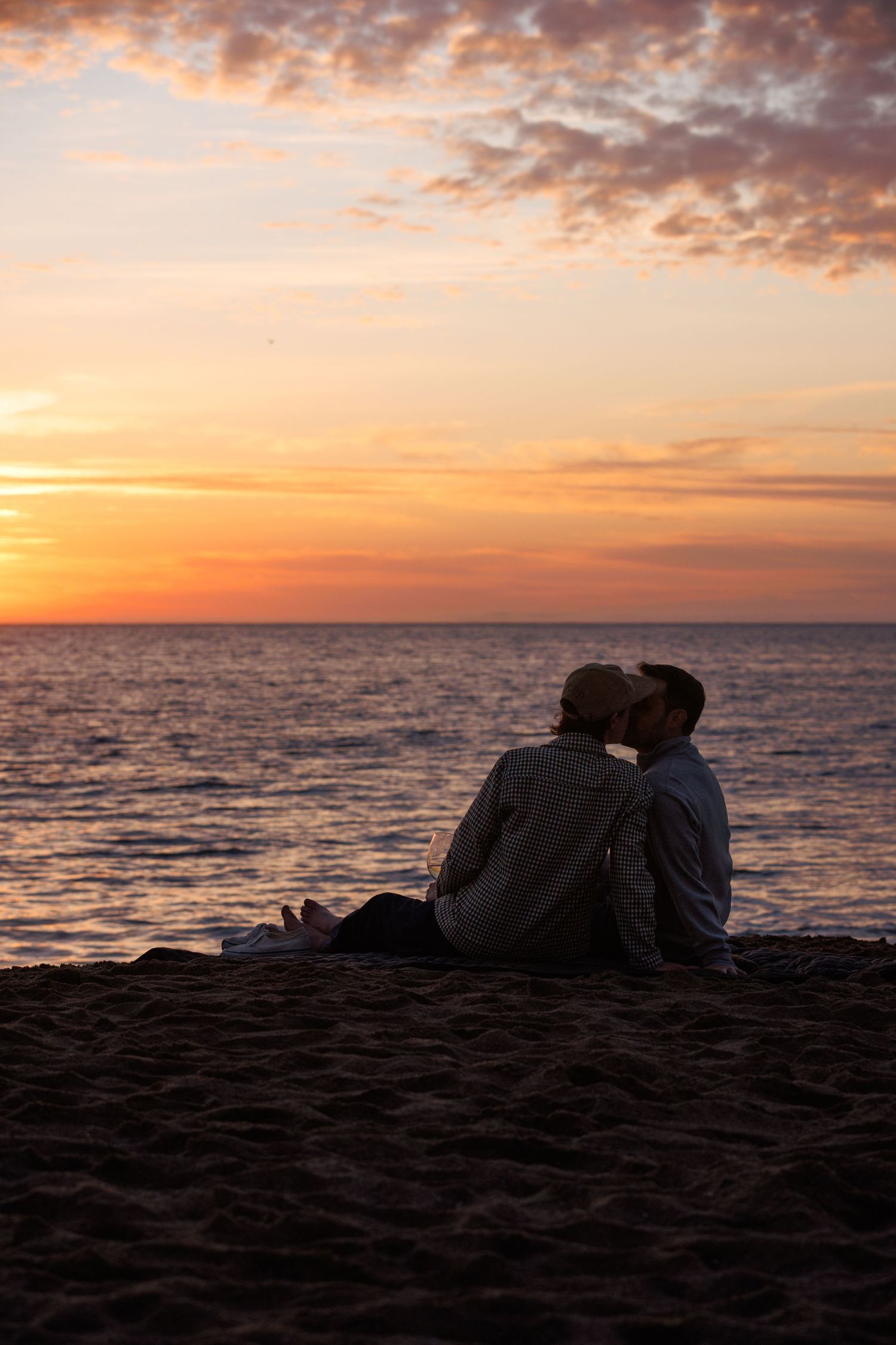Romantic couple silhouetted against a vibrant orange and pink sunset while sitting together on a sandy beach.