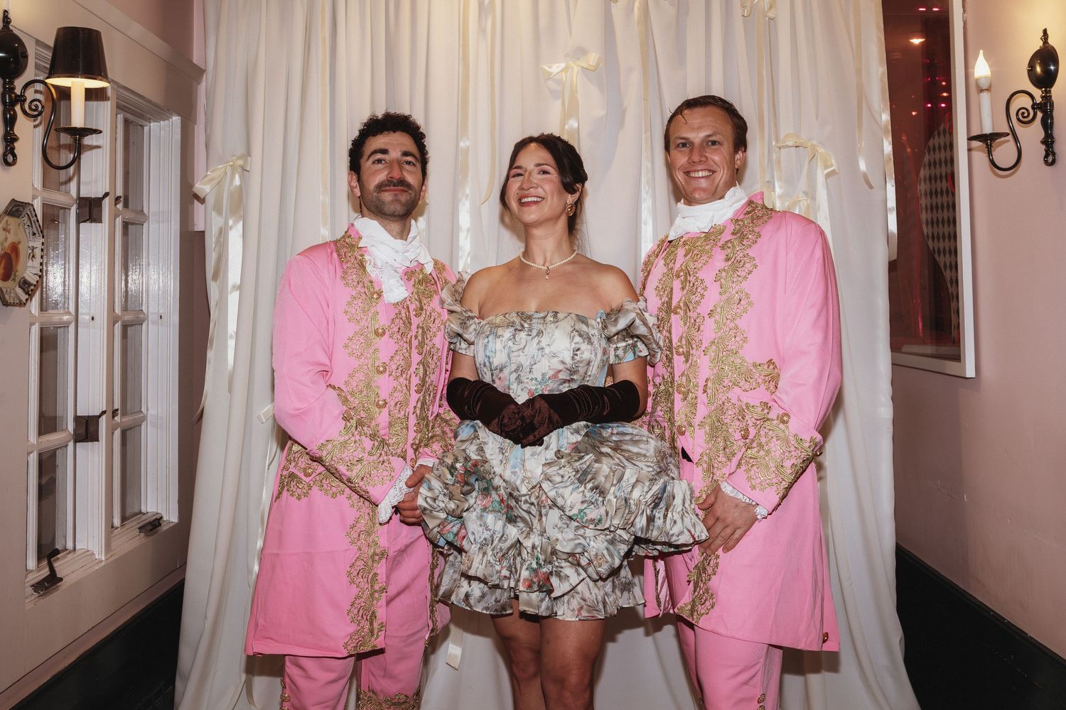 Two men in pink baroque jackets pose with a woman in a floral dress at an elegant wedding venue.