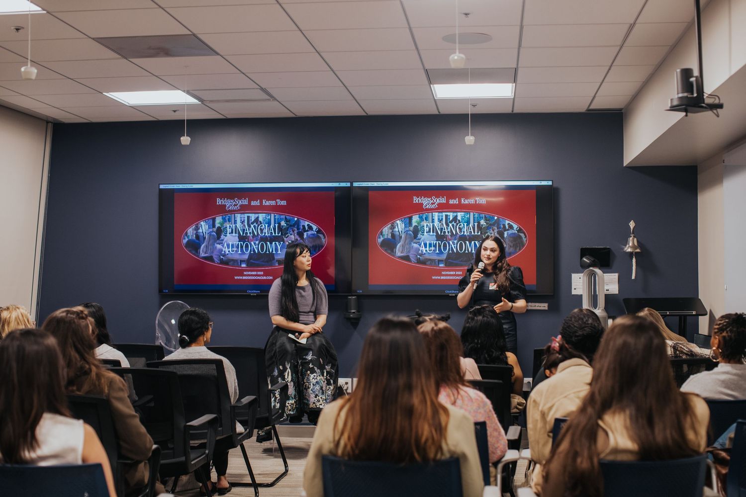 Two women speak on stage at a panel event with matching presentation screens behind them and an audience seated in rows.