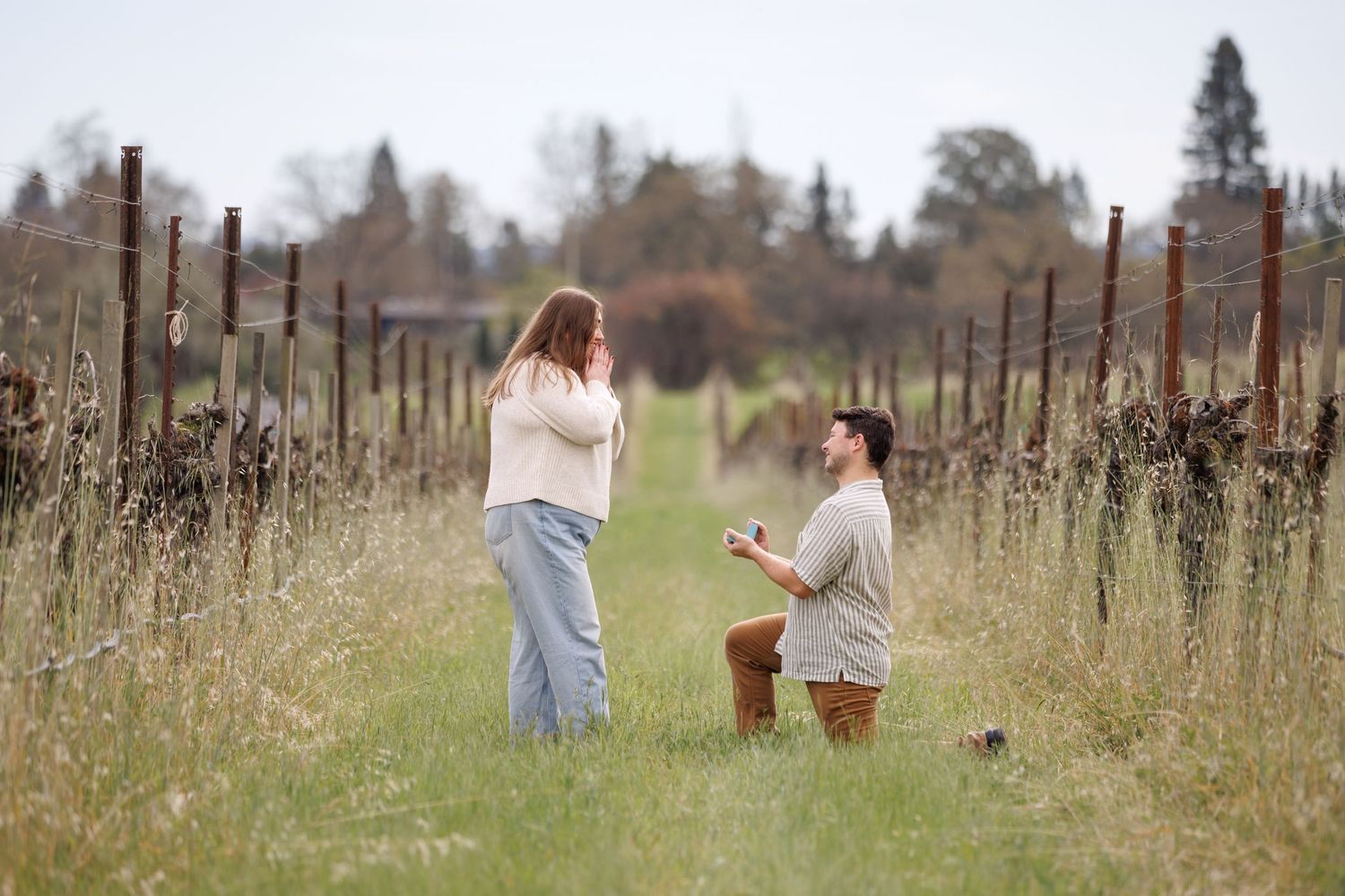 Man proposing marriage on one knee to surprised woman in a scenic vineyard field.