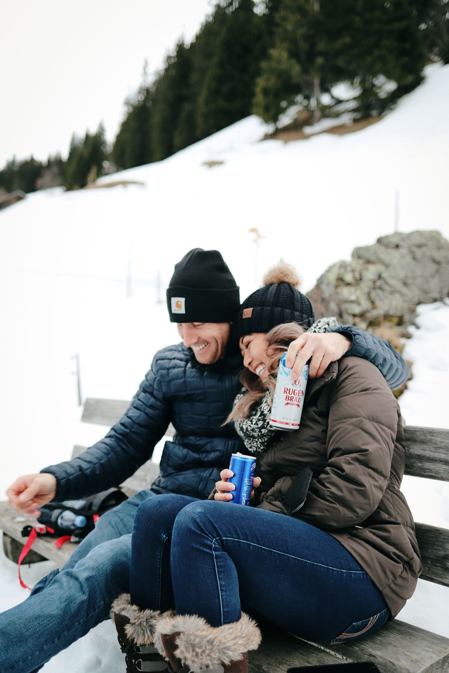 Couple in winter hats and jackets sitting on snowy mountain bench, laughing and sharing a Red Bull drink.