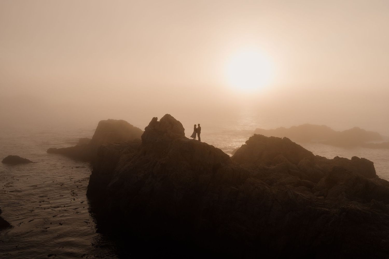 Two silhouetted figures stand on rocky coastal cliffs during a misty golden sunset over the ocean.