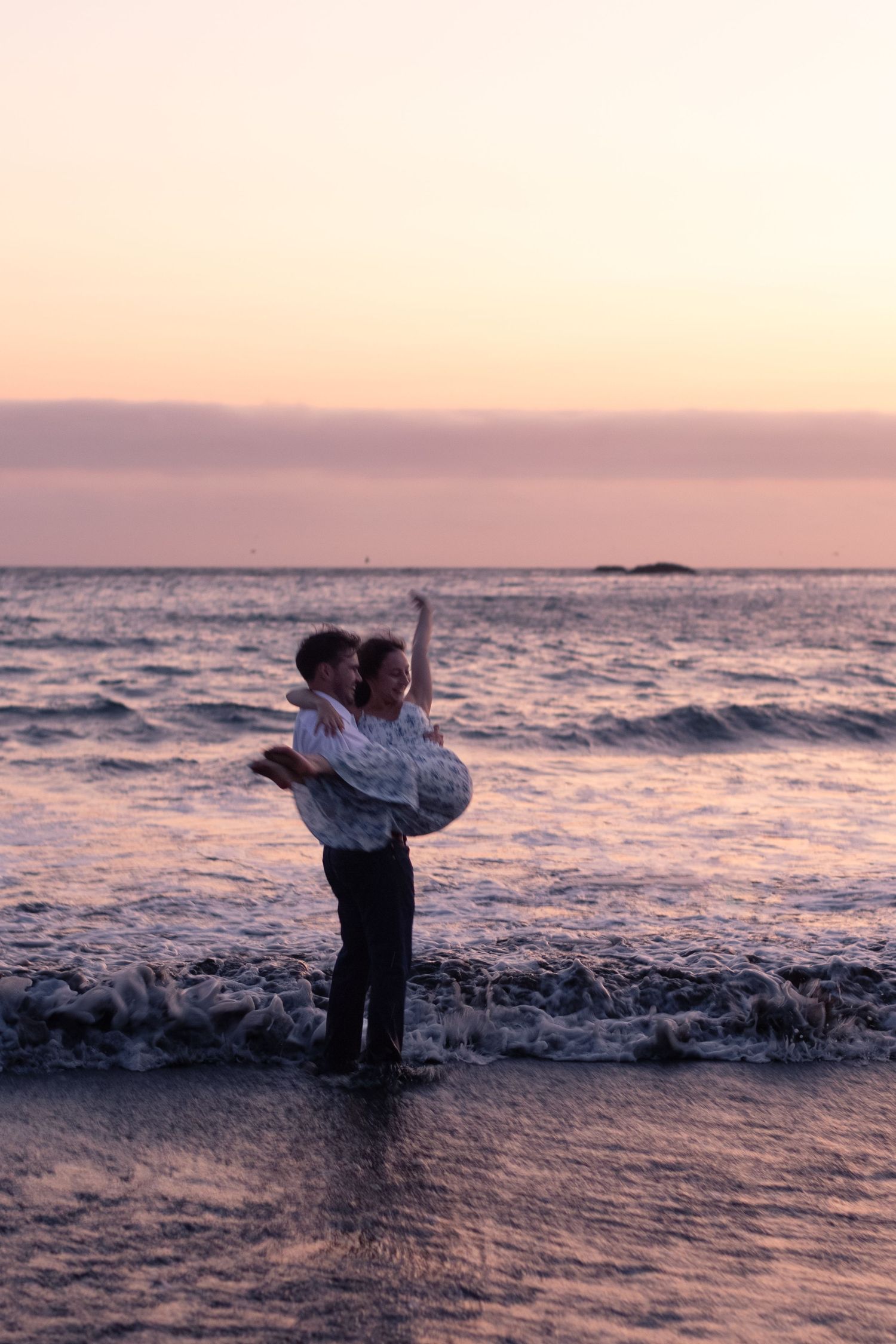 Couple embracing on a beach at sunset, waves washing ashore in a romantic golden-pink twilight scene.