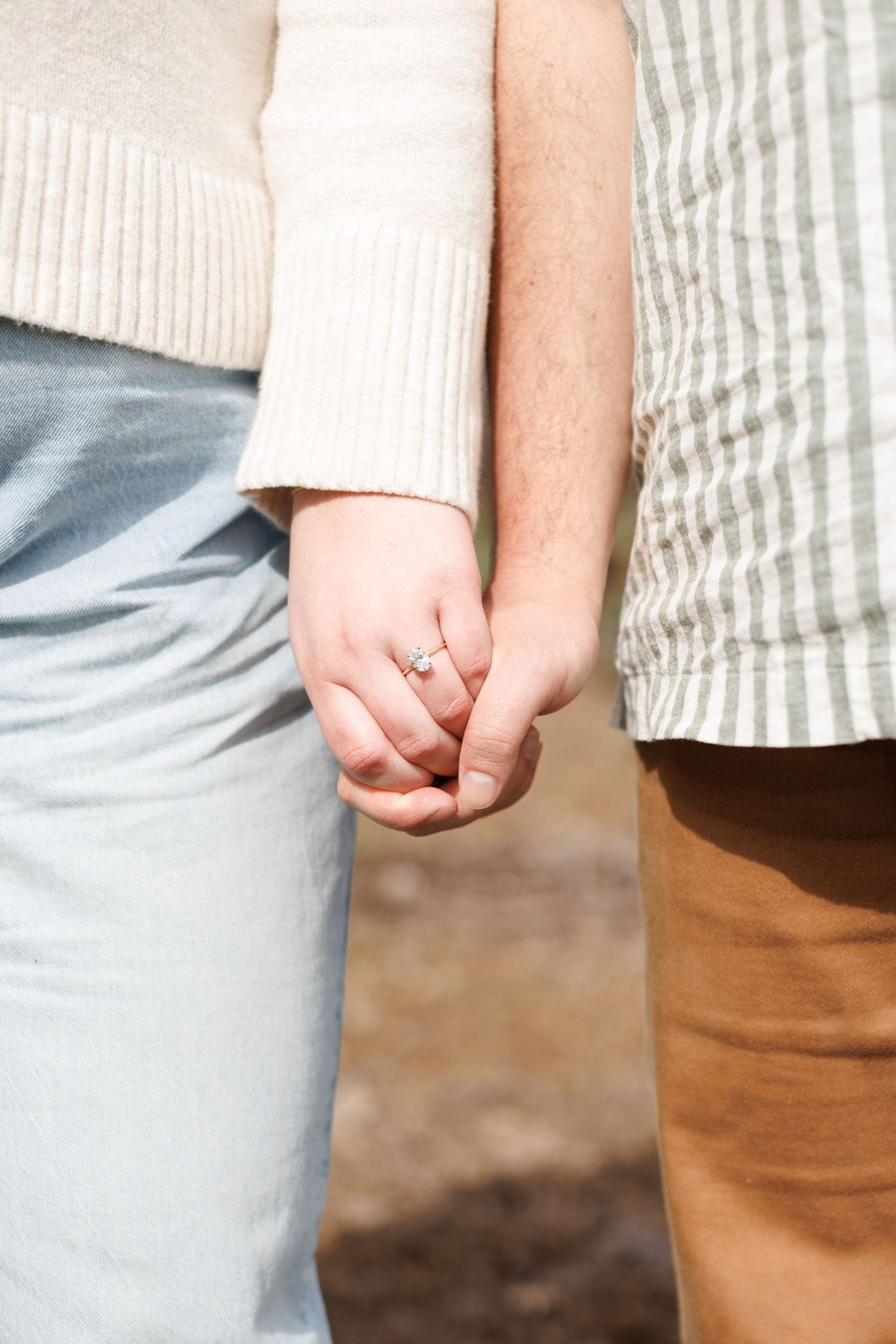 Close-up of a couple holding hands, woman wearing an engagement ring, dressed in casual sweater and skirt.
