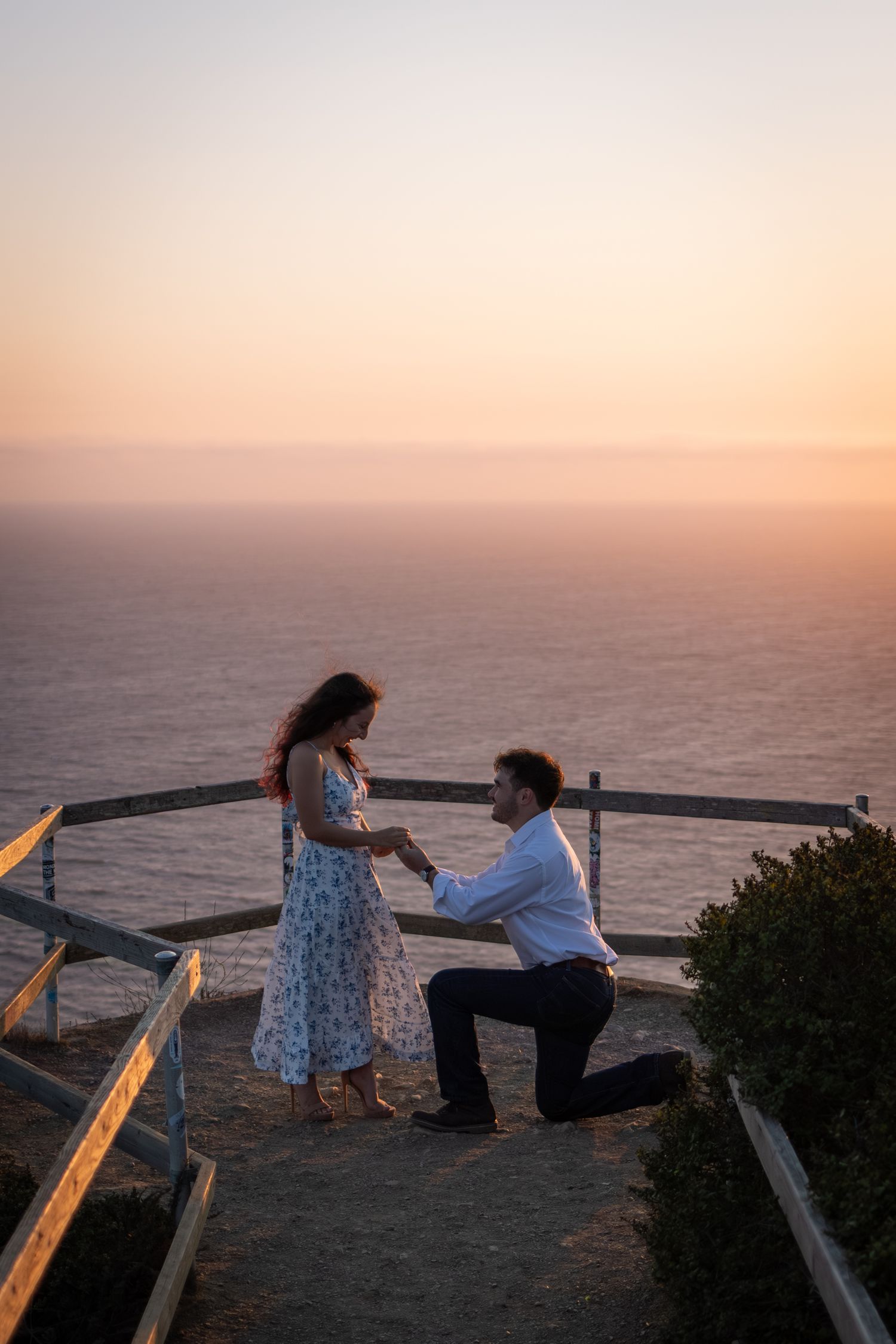 Man proposes to woman on one knee at scenic ocean overlook during golden sunset.