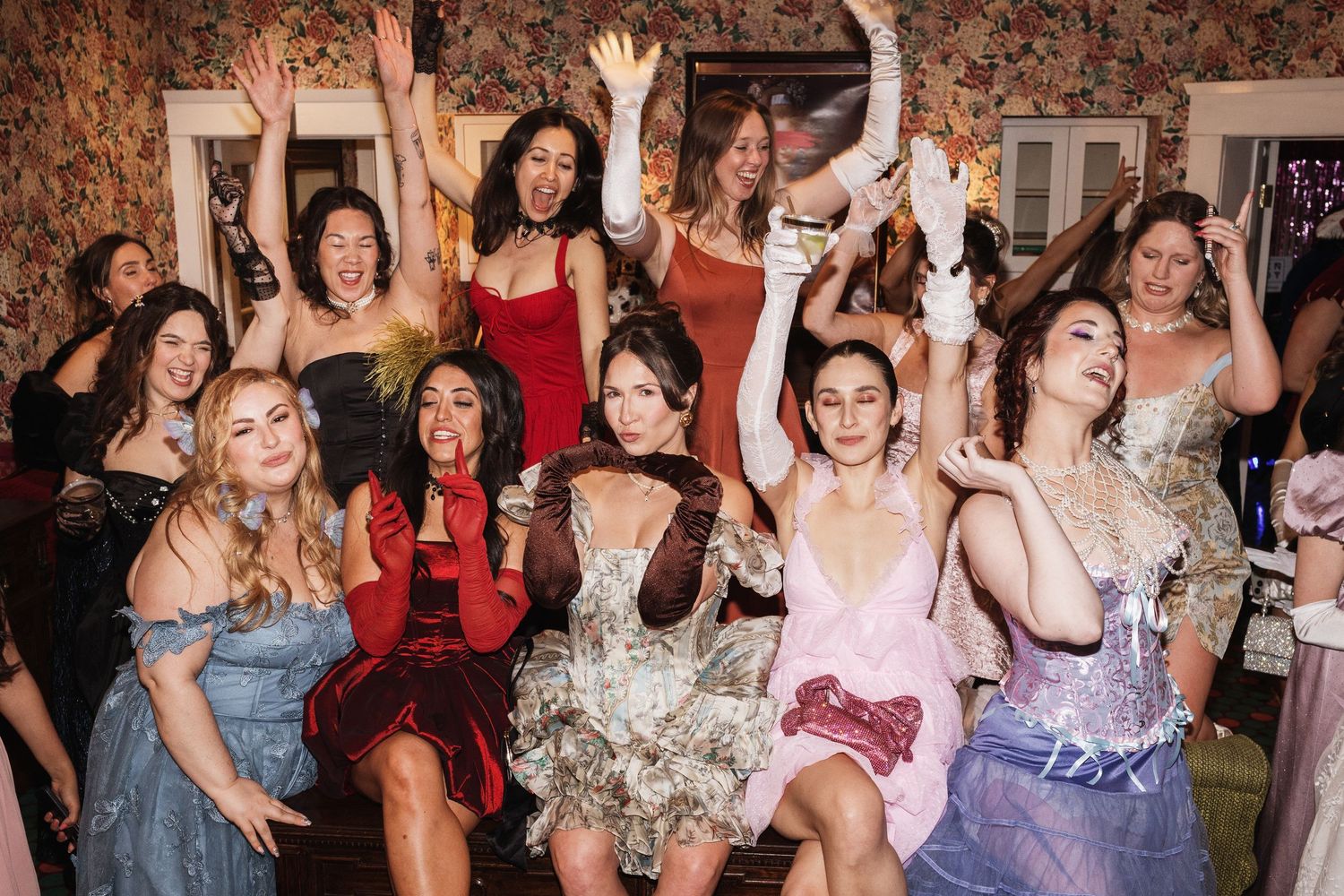Group of women in elegant dresses celebrating at a party, laughing and raising their arms in joy.