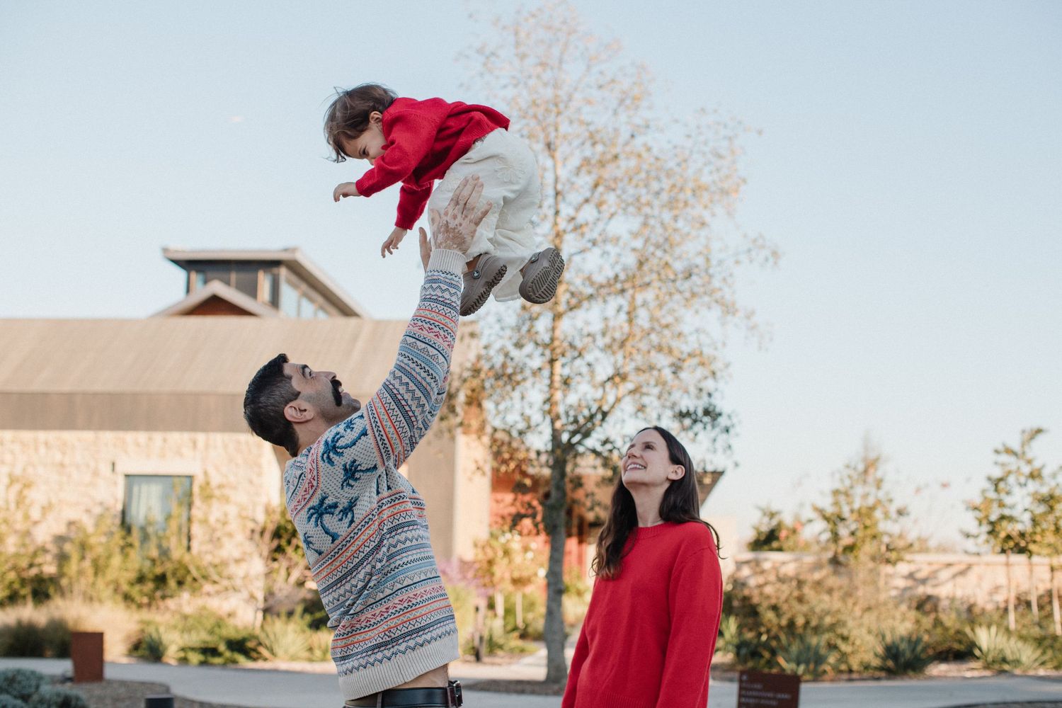 A parent lifts a child wearing red into the air while another person watches with joy in a residential setting.