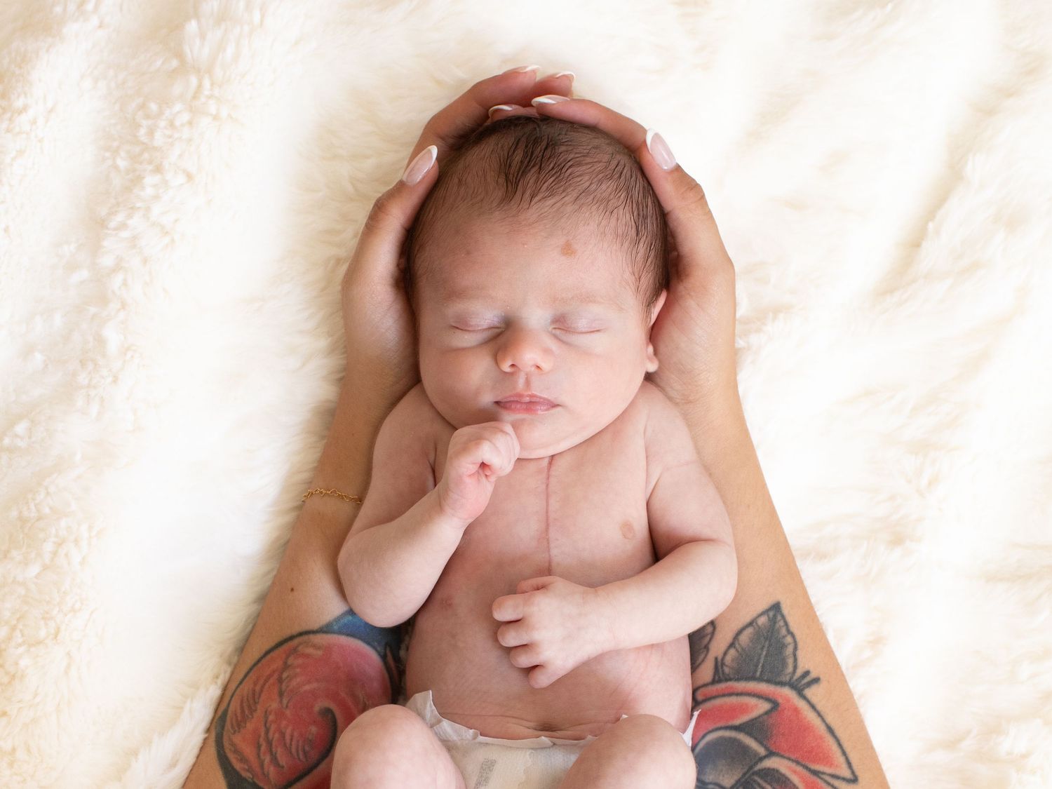 Sleeping newborn baby cradled in tattooed parent's hands on a soft cream blanket.