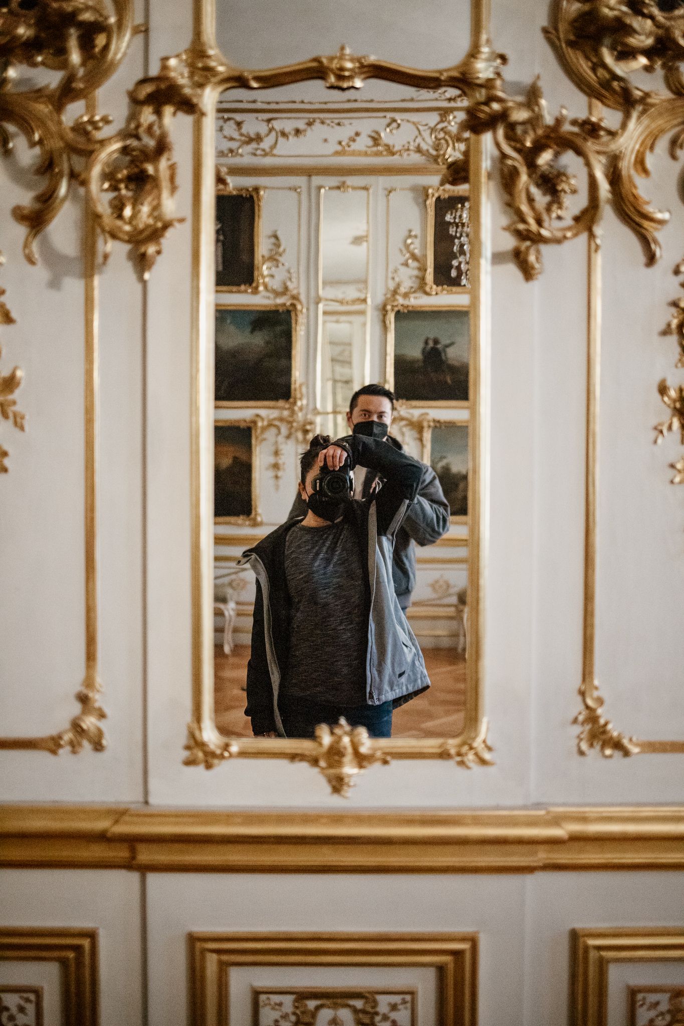Mirror selfie taken in an ornate baroque-style room with gilded decorative moldings and frames on white walls.