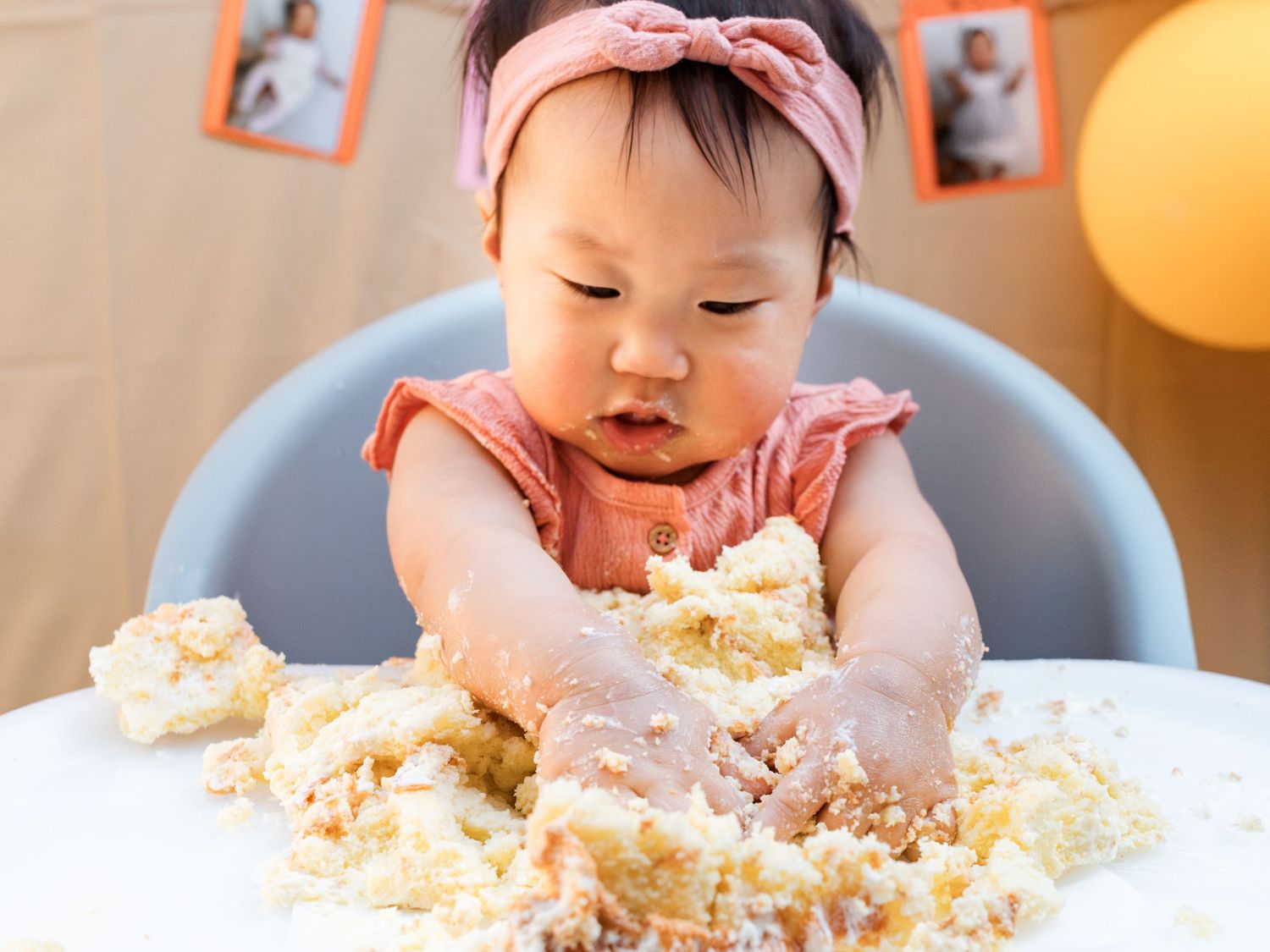 Baby girl in pink bow smashes first birthday cake in high chair at cake smash celebration.
