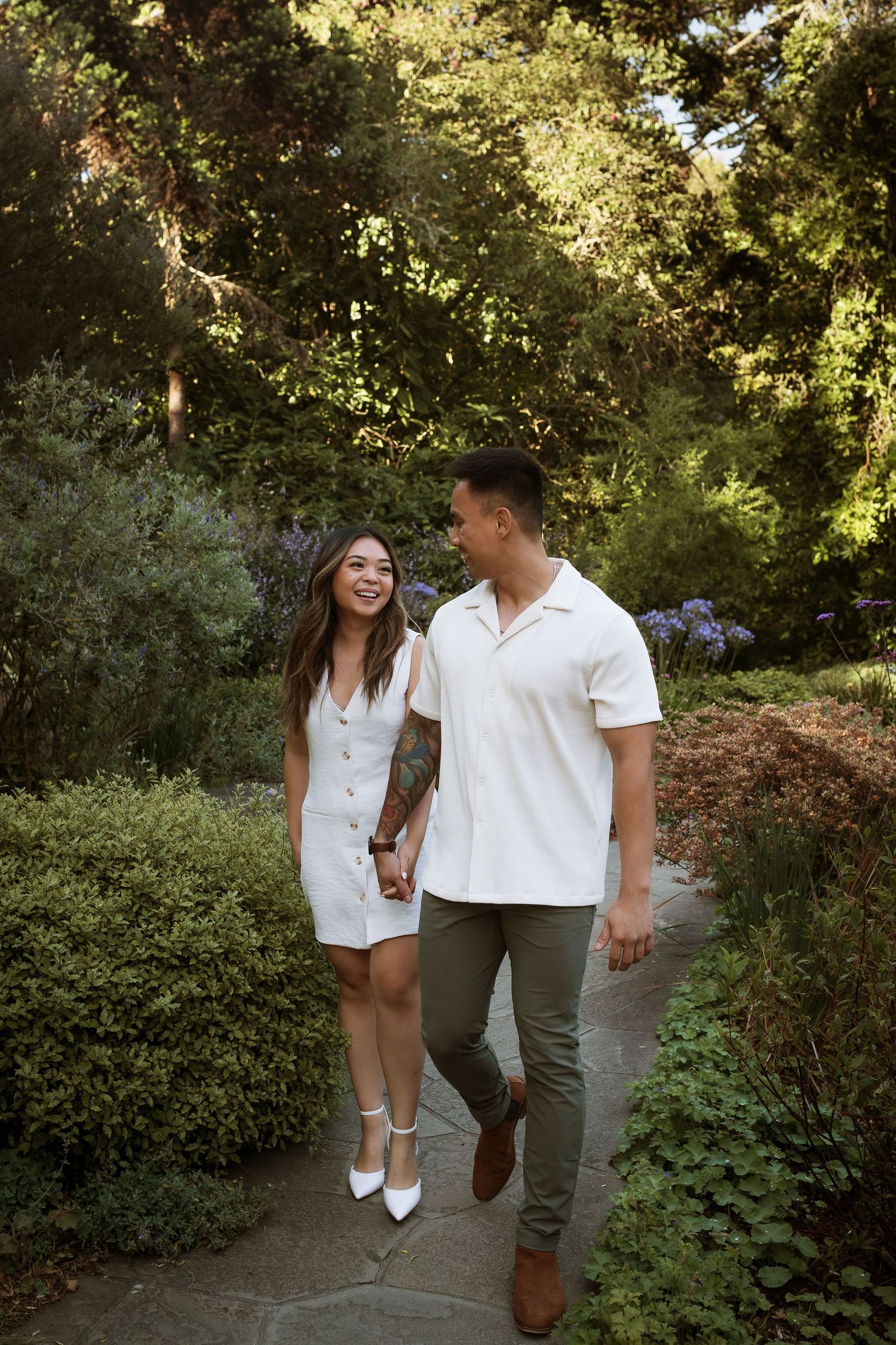 Couple holding hands and smiling while walking along a garden path surrounded by lush green trees and shrubs.