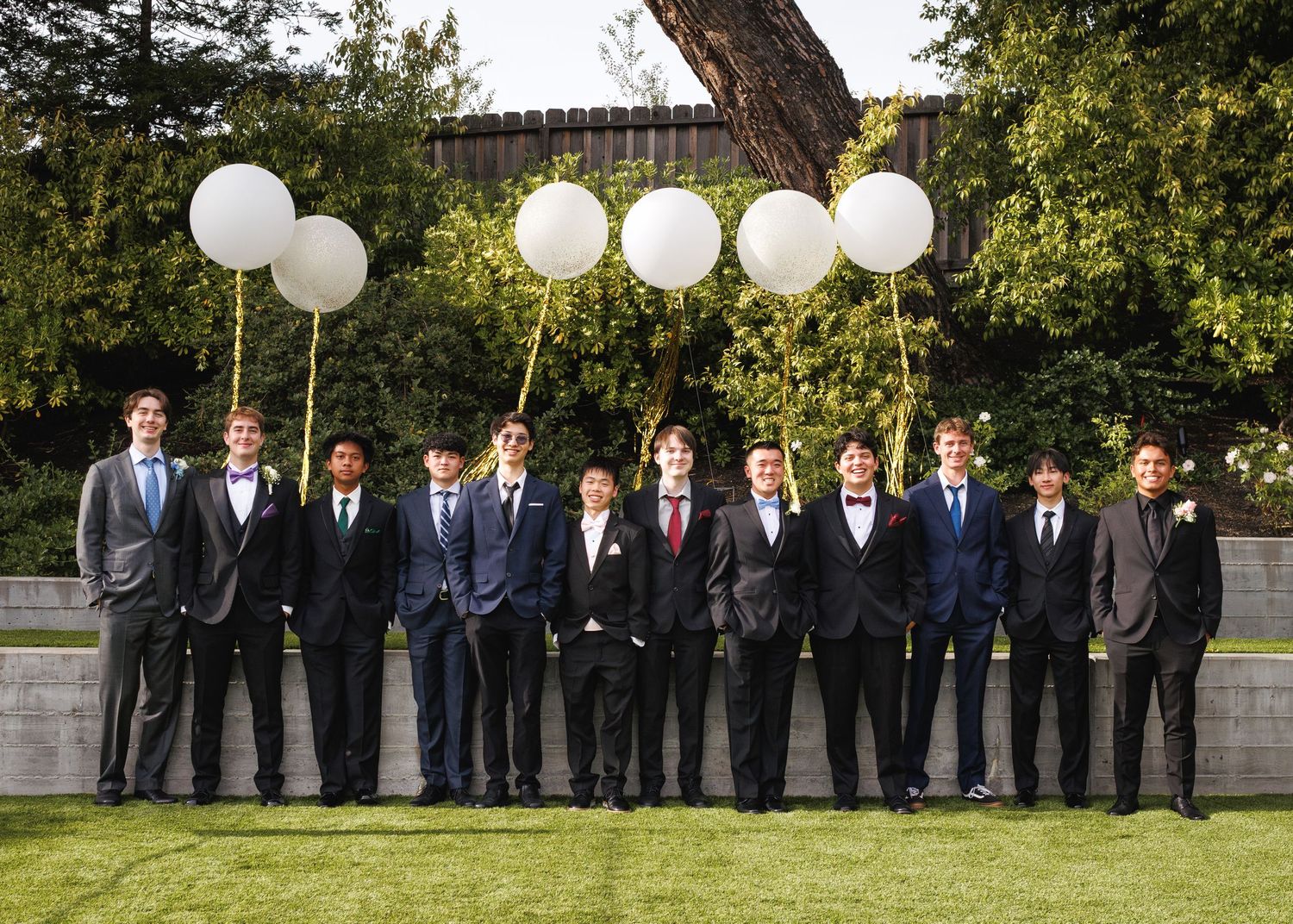A group of young men in formal suits pose outdoors with large white balloons at what appears to be a prom event.