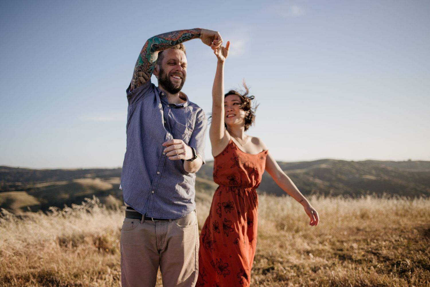 A couple dances together in casual attire on a hilltop meadow during golden hour with mountains in the background.