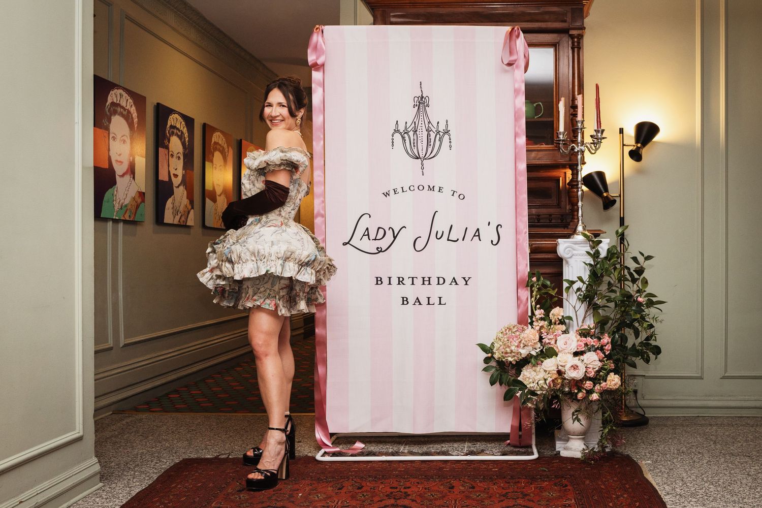 Woman in ruffled dress posing beside Lady Julia's Birthday Ball welcome sign with pink stripes and chandelier.