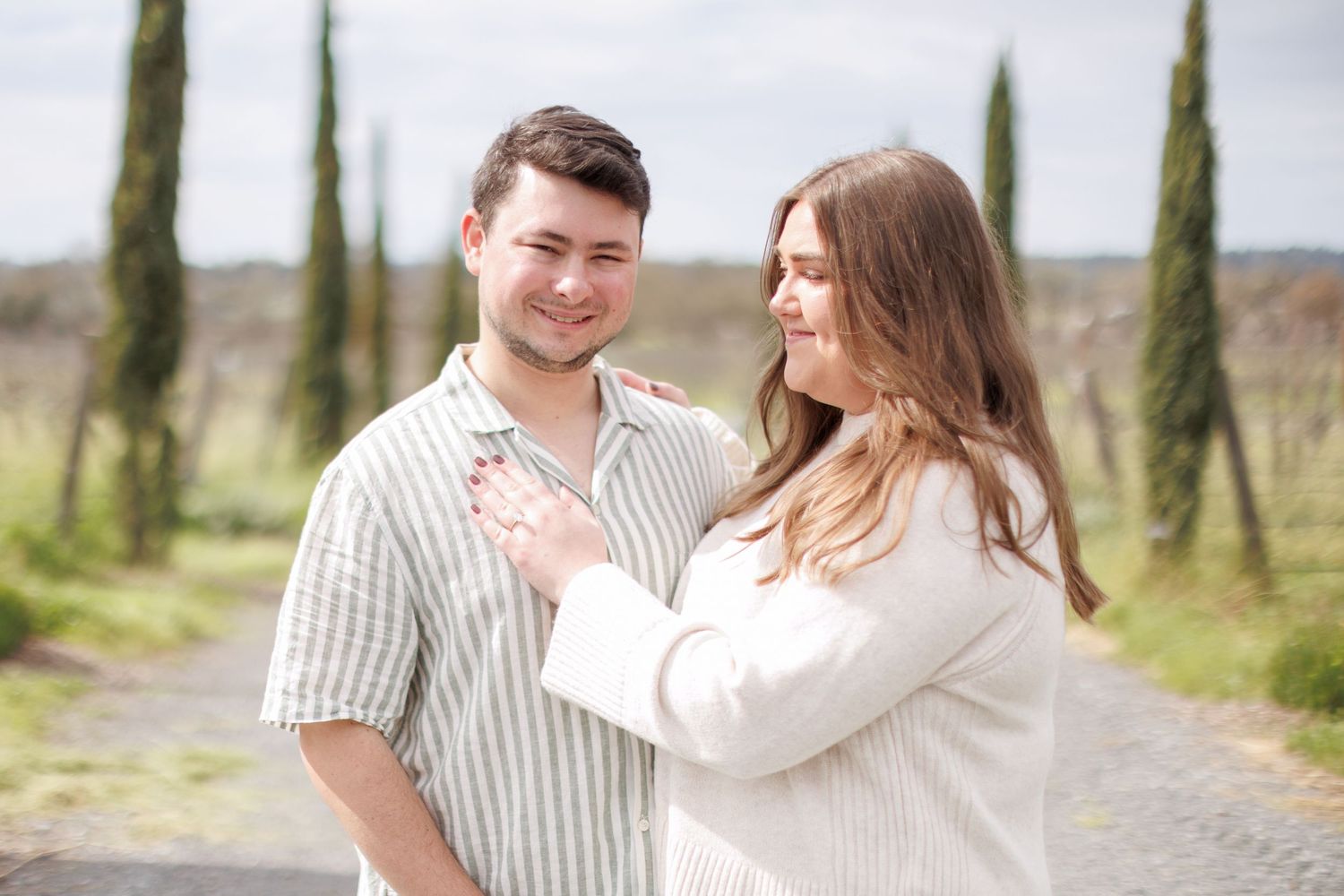 Couple posing in vineyard, woman in white sweater resting hand on mans chest, tall cypress trees in background.