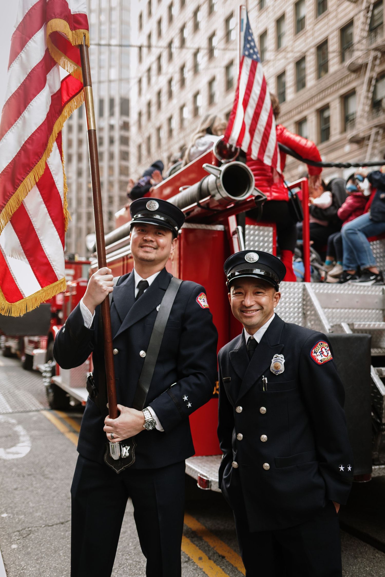 Two firefighters in dress uniforms smile beside a red fire truck during a parade, one holding an American flag.