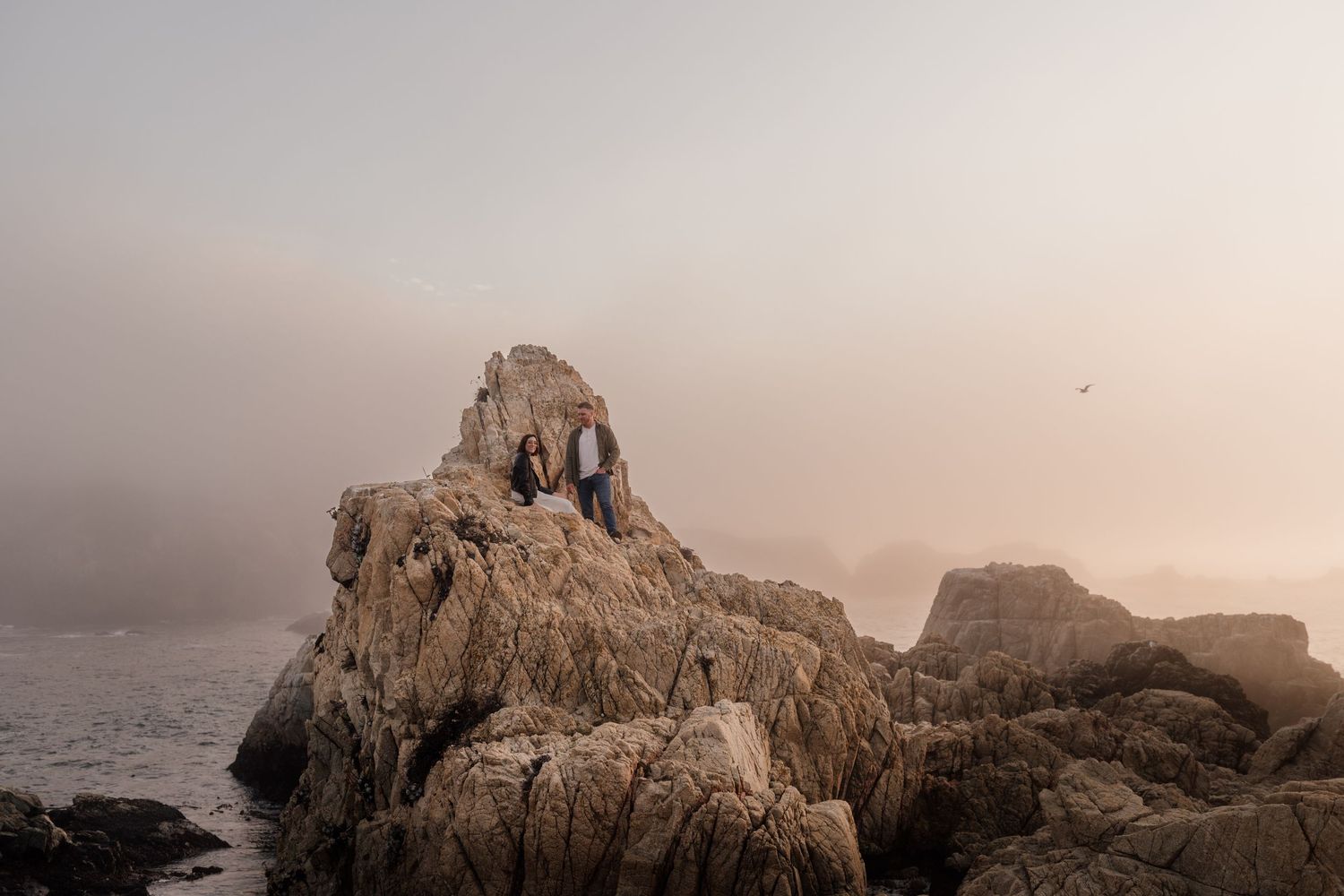 Couple sitting atop rugged coastal rocks shrouded in misty fog near the ocean at dusk.