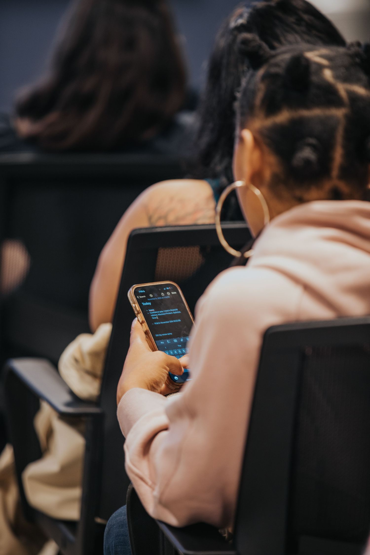 Young woman with hoop earrings using smartphone while seated, viewed from behind in casual setting.