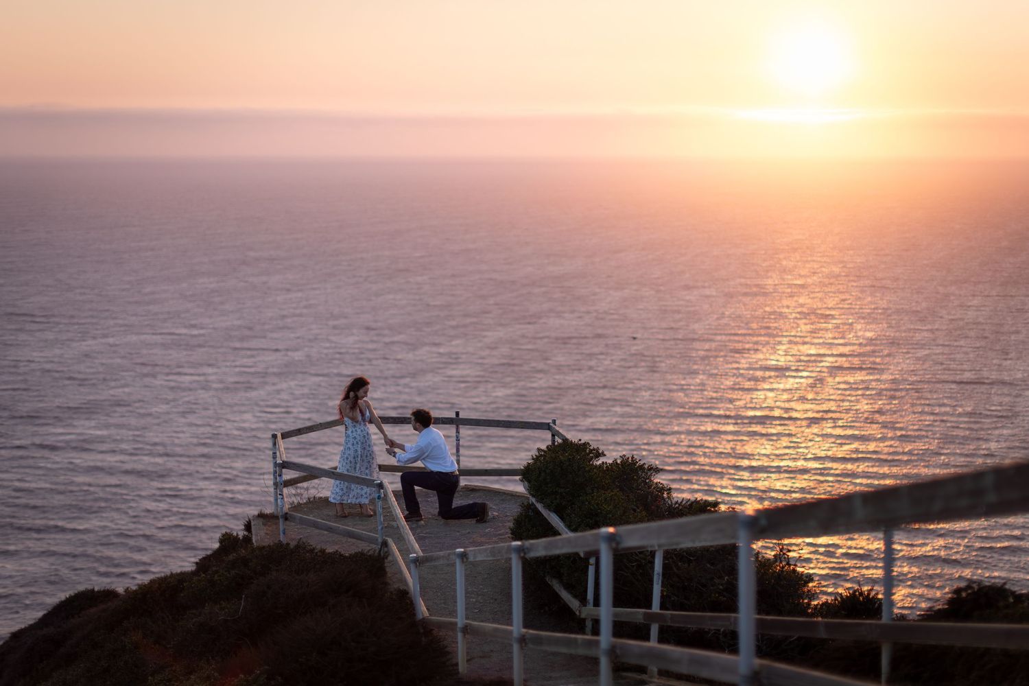 Couple sharing a romantic sunset proposal moment on a clifftop ocean overlook platform with golden sky.