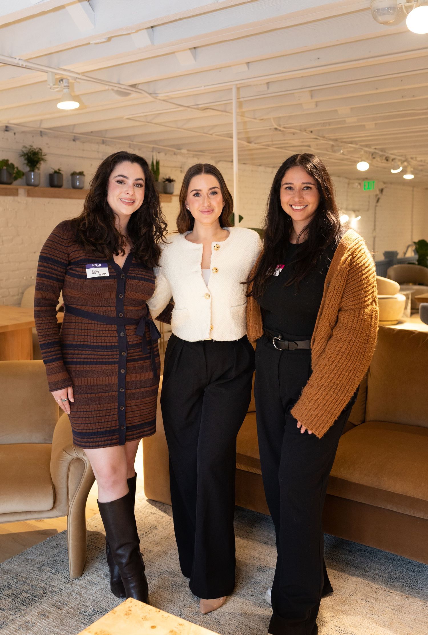 Three women smiling together at an indoor networking event, dressed in stylish outfits.