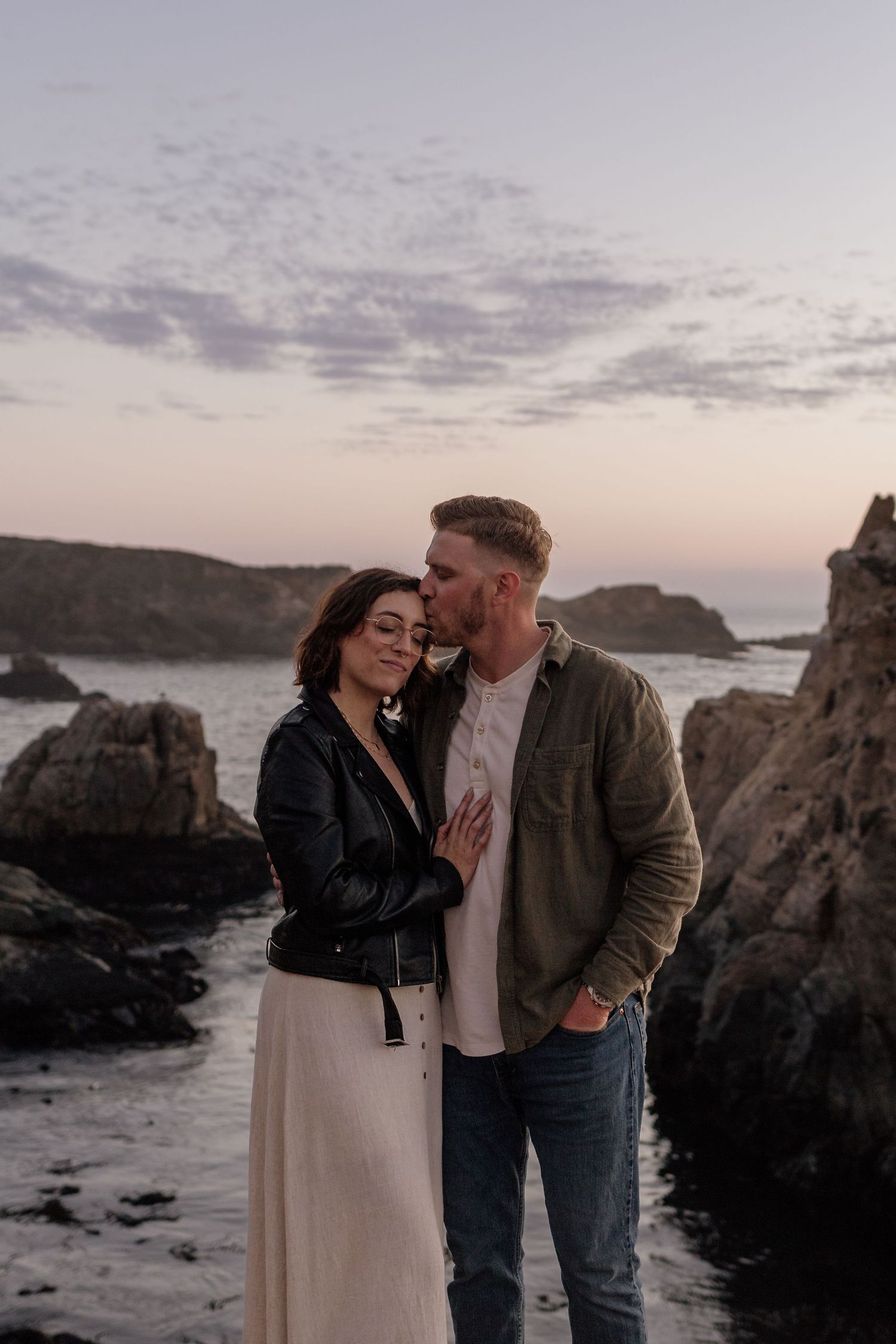Couple embracing at sunset on rocky Big Sur coastline, man kissing woman's forehead, wearing casual jackets.
