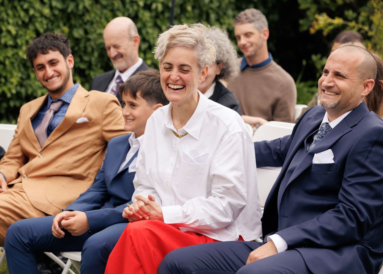 Group of smiling people seated outdoors at a garden event, dressed in elegant attire, sharing a joyful moment.