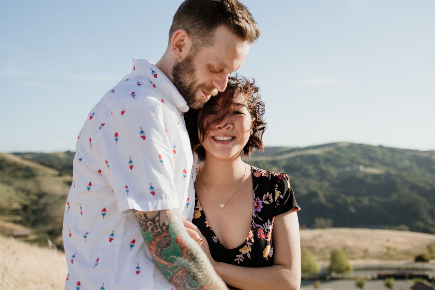 A smiling couple embraces outdoors with rolling green hills and blue sky in the background on a sunny day.