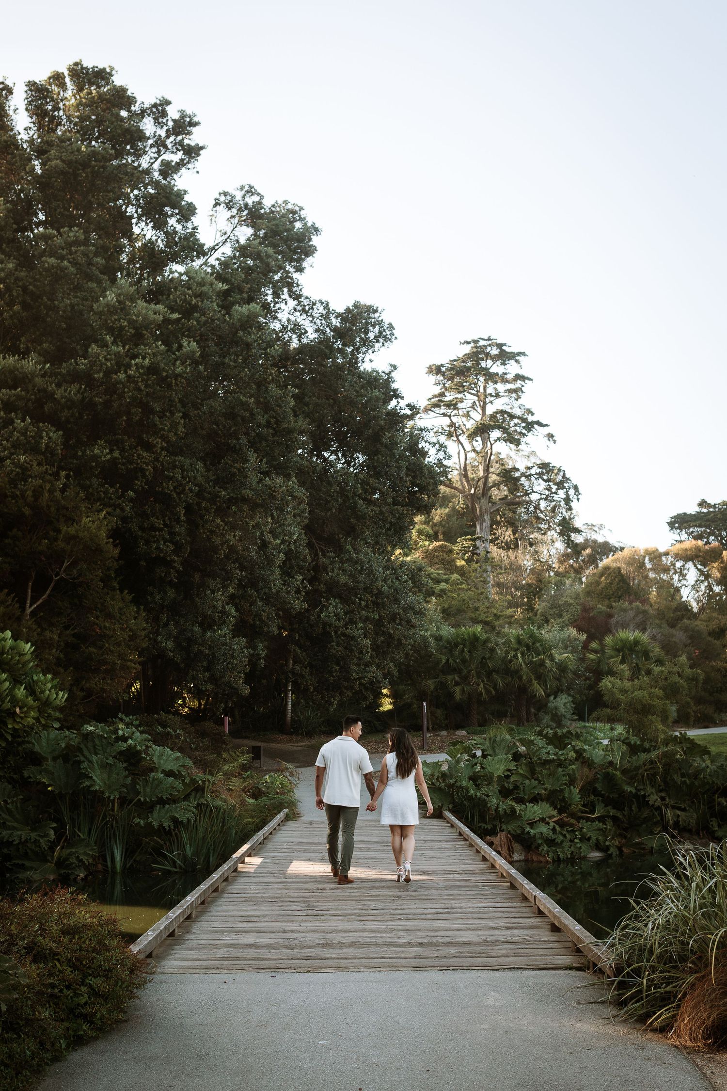 Couple walking hand in hand along a wooden boardwalk path surrounded by lush green trees in a park.