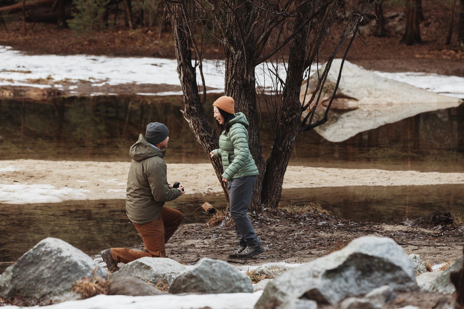 Two hikers in winter jackets stand near a bare tree by a snowy riverbank in a forest setting.