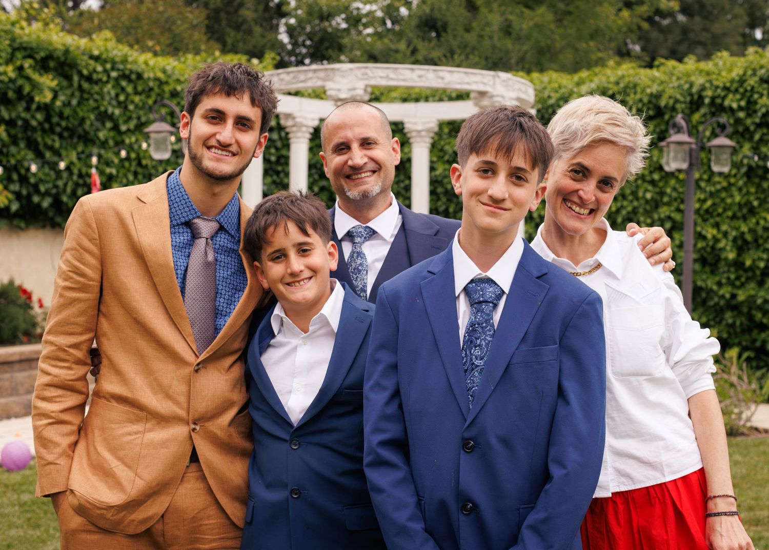 Family of five posing together outdoors at a formal event, wearing suits and dressy attire near a garden gazebo.