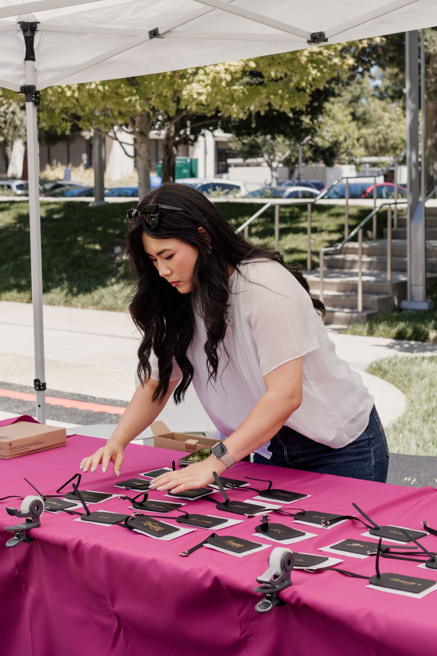 Woman arranging black cards on a pink tablecloth under a white canopy tent at an outdoor event.