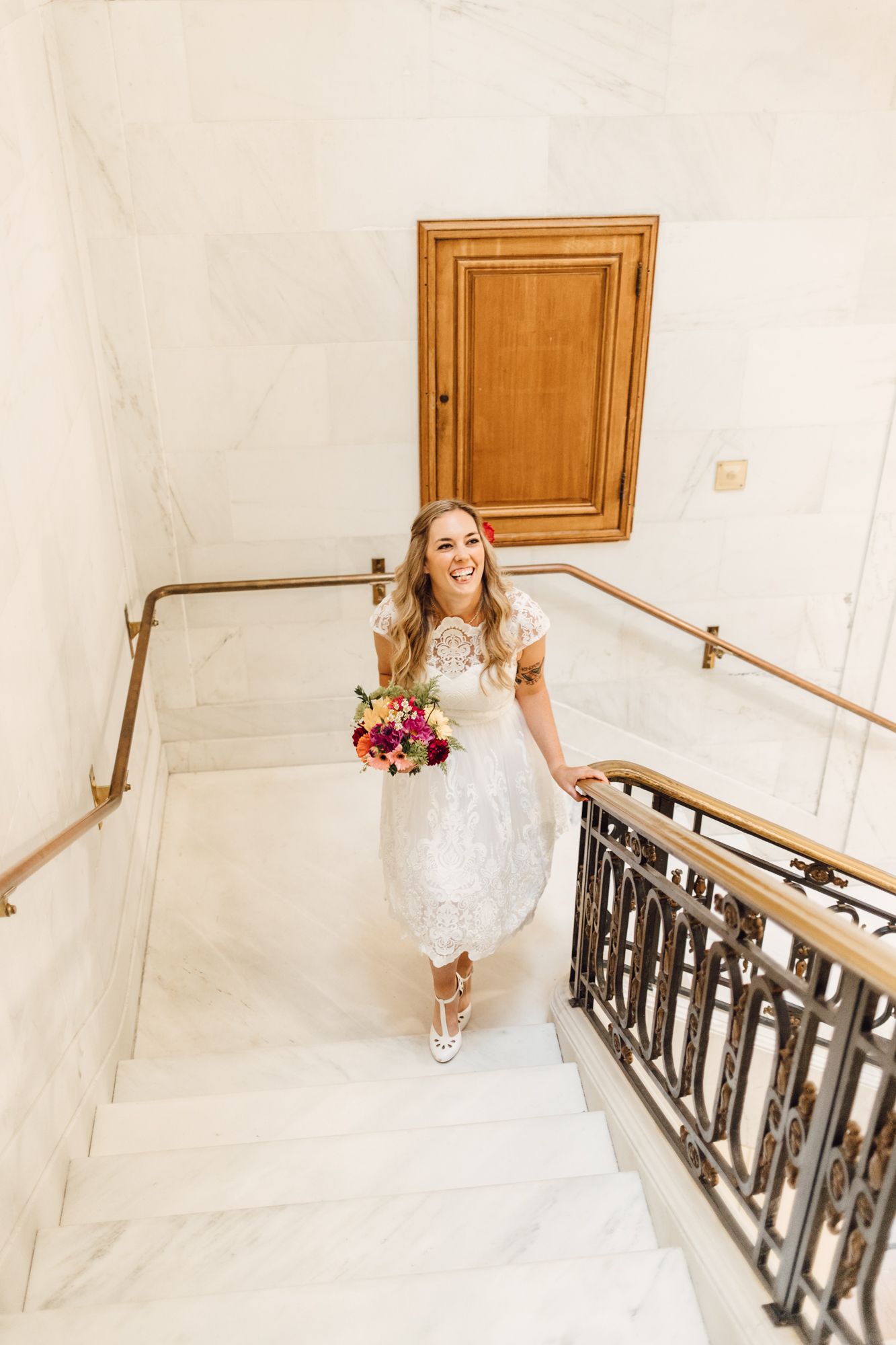 Smiling bride in white lace dress holding colorful bouquet walking up marble staircase.